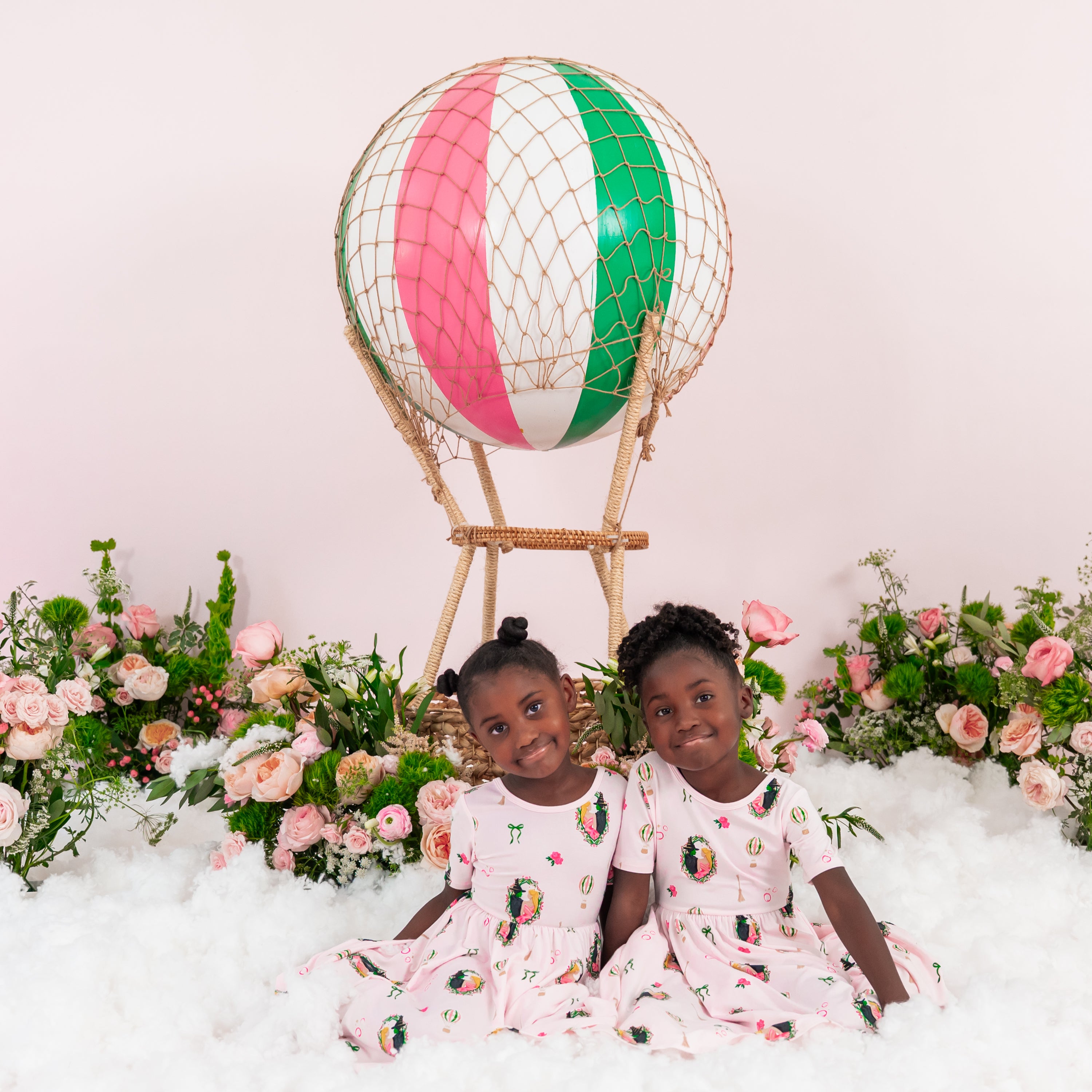 Two young girls sitting in front of a prop hot air balloon and flowers both wearing the Twirl Dress in Wicked