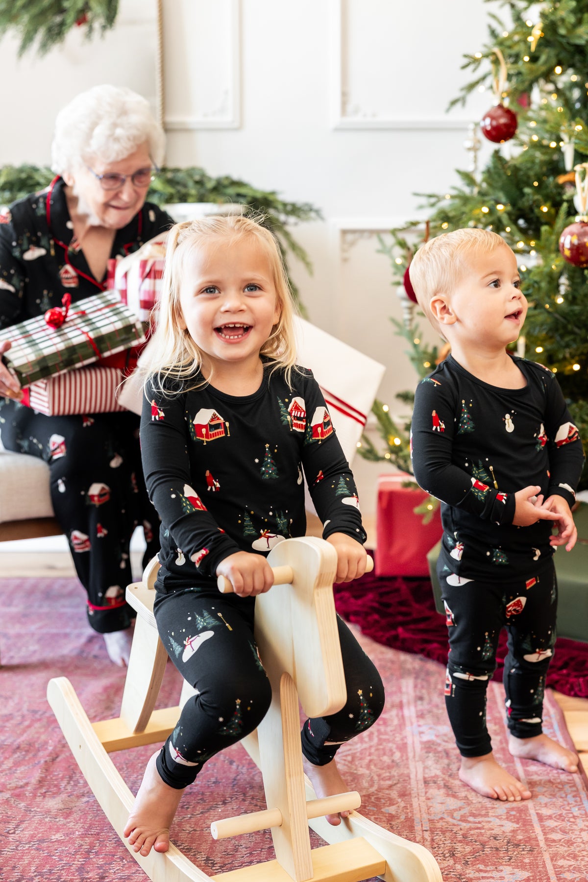 Two toddlers wearing matching black Christmas pajamas with holiday prints are in a living room by a Christmas tree. The girl sits on a rocking horse, and an older woman in matching pajamas holds wrapped gifts in the background.