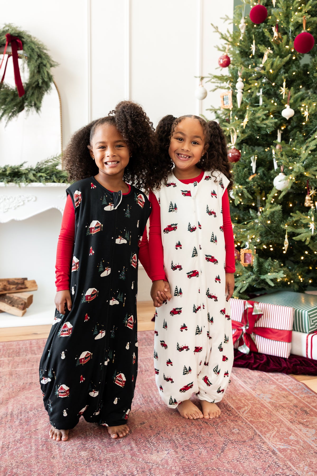 Two young girls standing barefoot in front of a decorated Christmas tree and fireplace, wearing matching holiday sleep sack walkers. One sleeper is black with red and white holiday motifs, and the other is white with red and green holiday motifs, both with red long-sleeve undershirts visible.