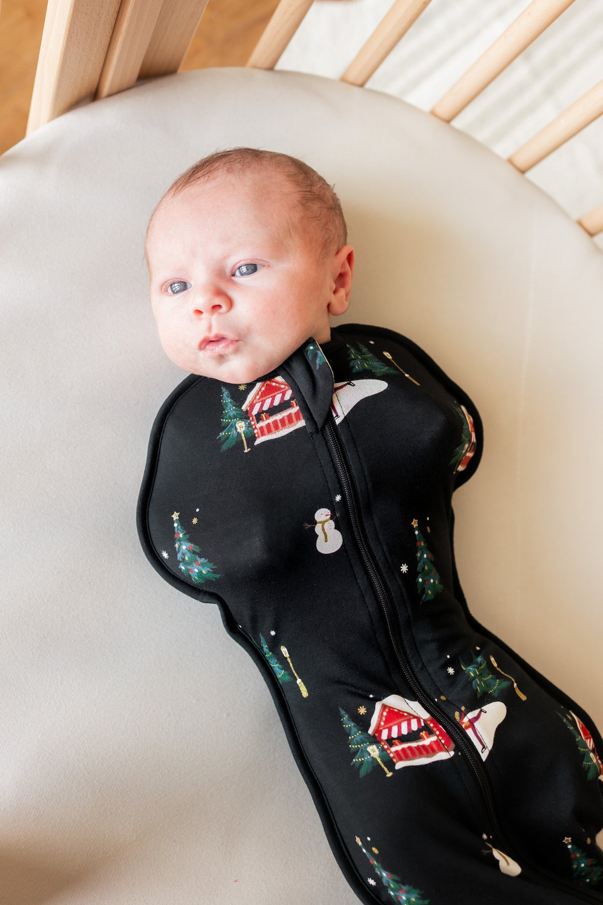 A baby lying on their back in a cream-colored changing crib, wearing a black footed sleep sack with a Christmas print of snowmen, trees, and houses.
