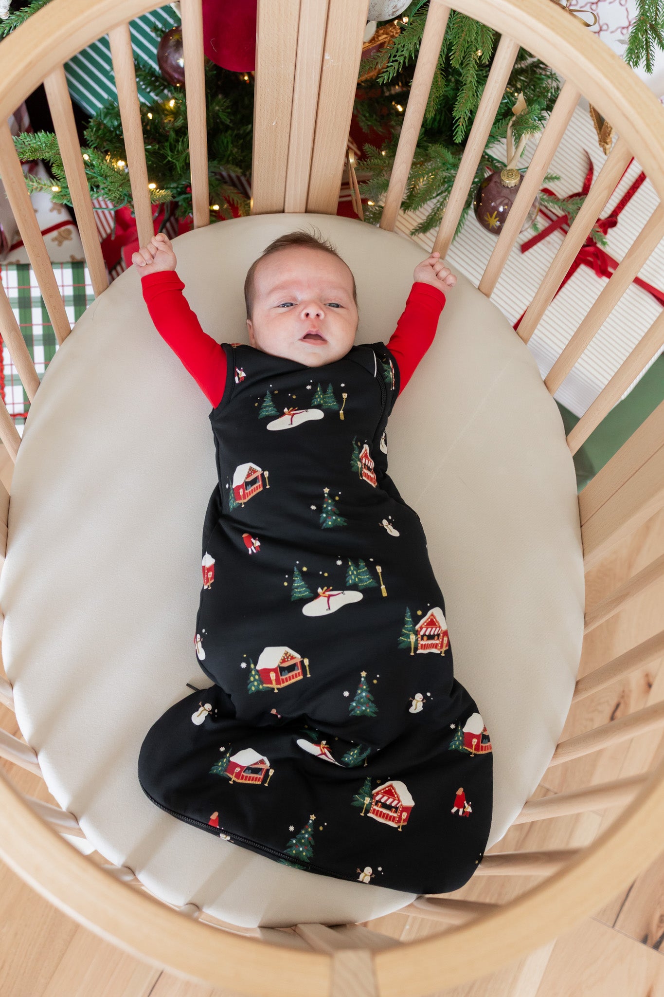 A baby lying in a wooden crib, wrapped in a black swaddle blanket with a Christmas village print. The baby is wearing red long-sleeve socks, and the crib has a cream mattress.
