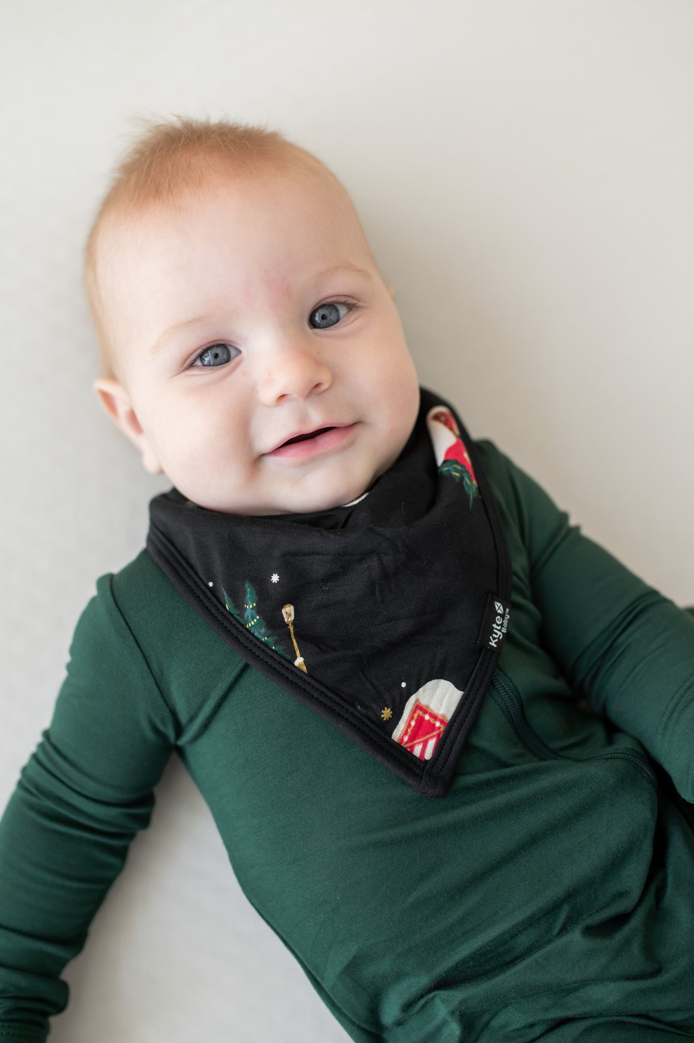 Baby wearing bib in winter wonderland print over pine zippered footie on a beige background