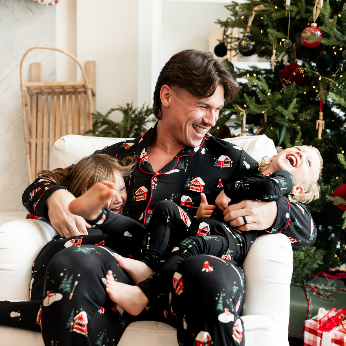 Father and 2 kids wearing black pajamas with winter wonderland motifs with christmas tree in background