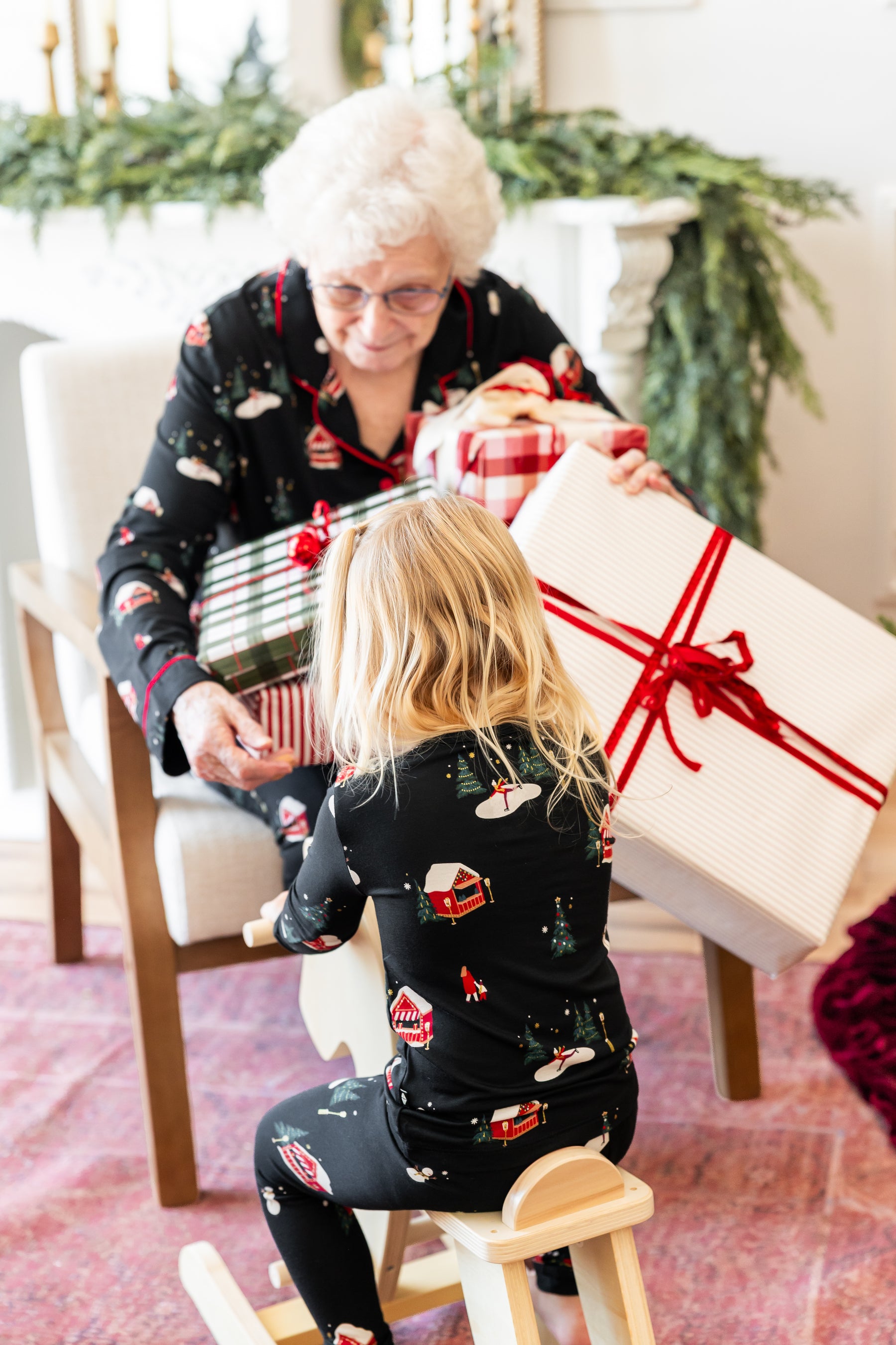 A woman and a young child wearing matching black Christmas pajamas with a holiday print are sitting in a cozy living room, sharing large wrapped gift boxes with red ribbons.