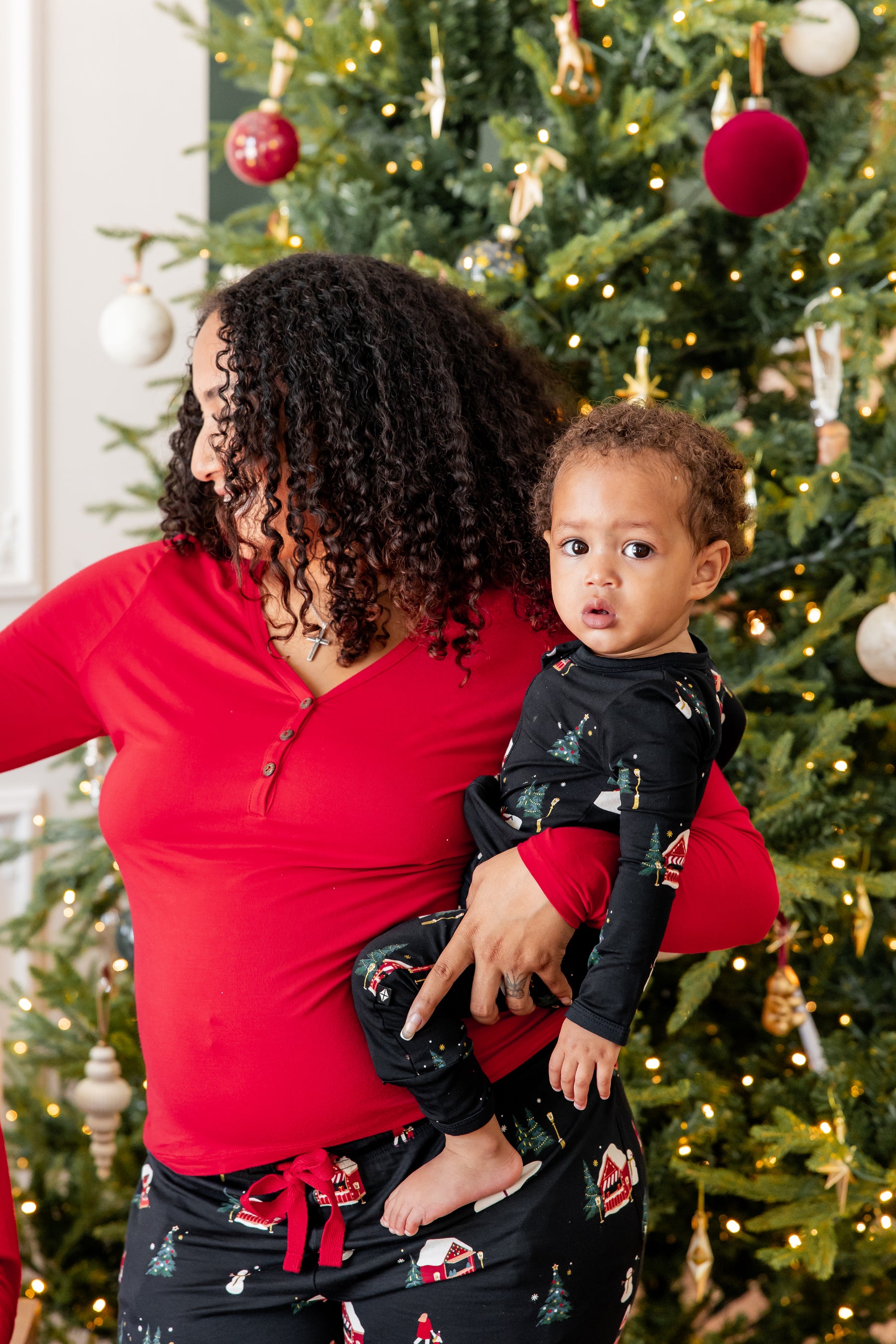 A woman and a toddler wearing matching Christmas-themed pajamas, standing in front of a decorated Christmas tree. The woman wears a red long-sleeve top and black patterned pants, while the toddler wears a black long-sleeve top and matching black pants.