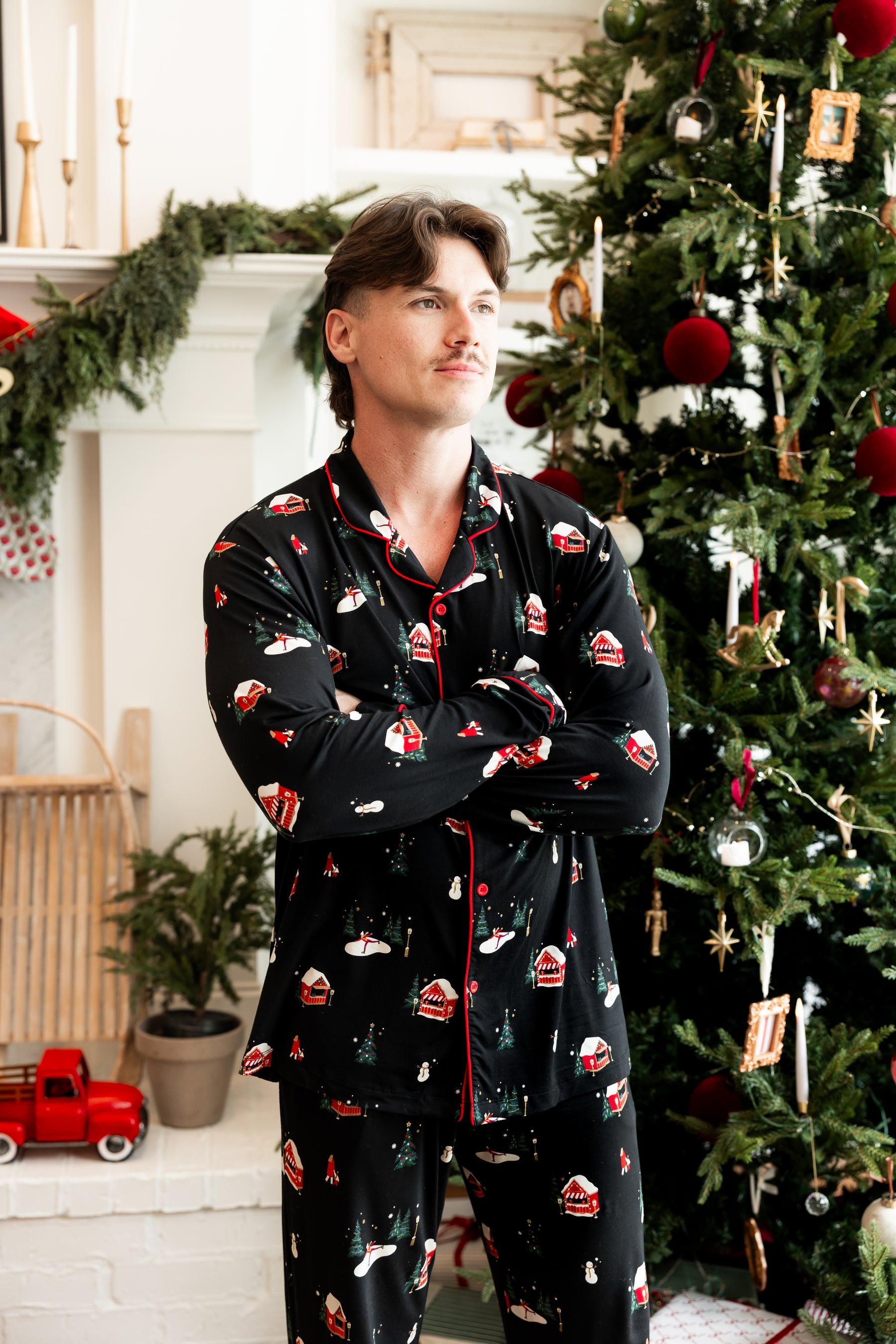 A man wearing a black long-sleeve pajama set with a red and white Christmas print stands in front of a decorated Christmas tree and fireplace mantel.