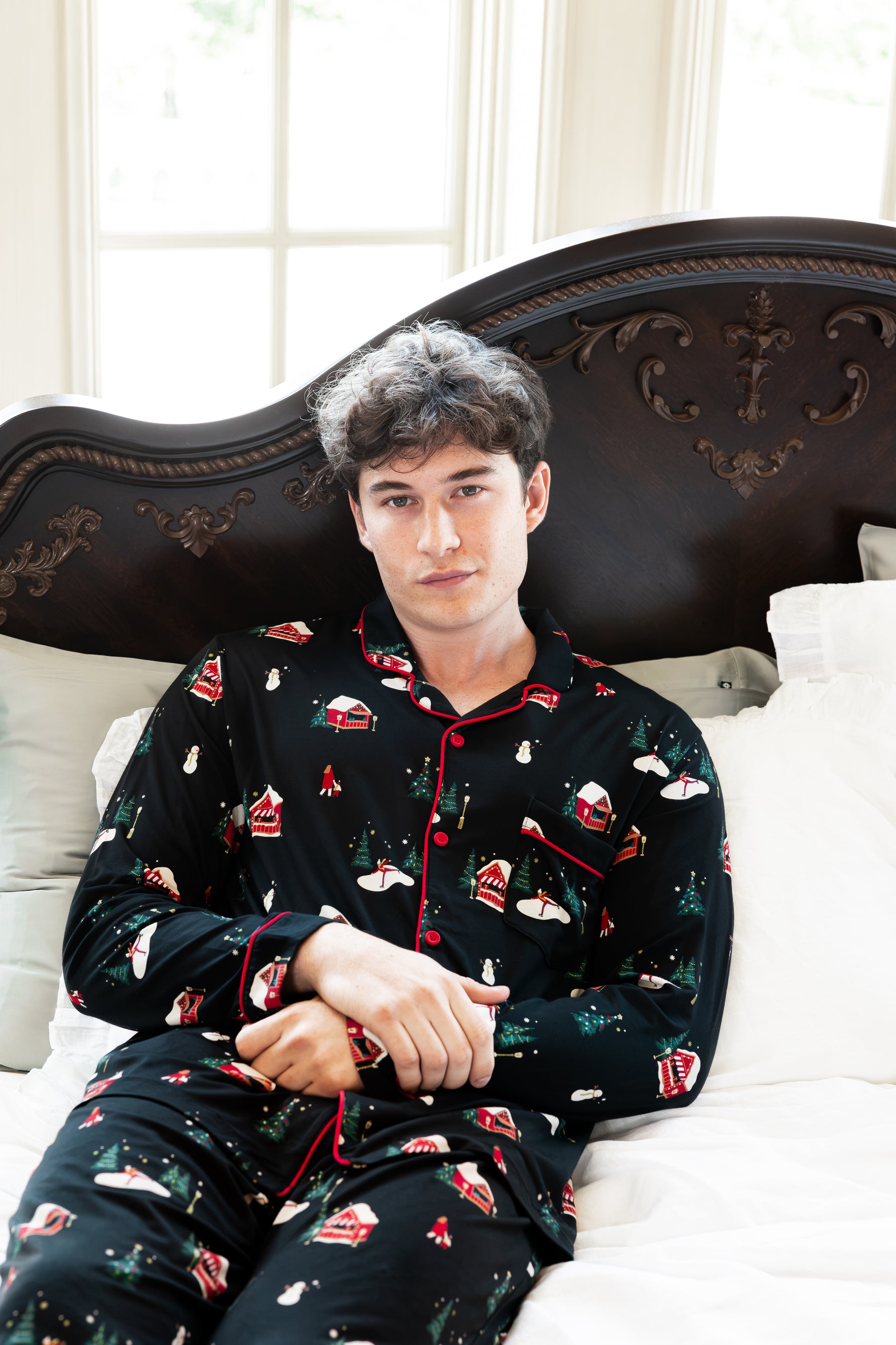 A man wearing a black long-sleeve pajama set with a red and green Christmas print, sitting on a bed with white pillows and a dark wooden headboard.