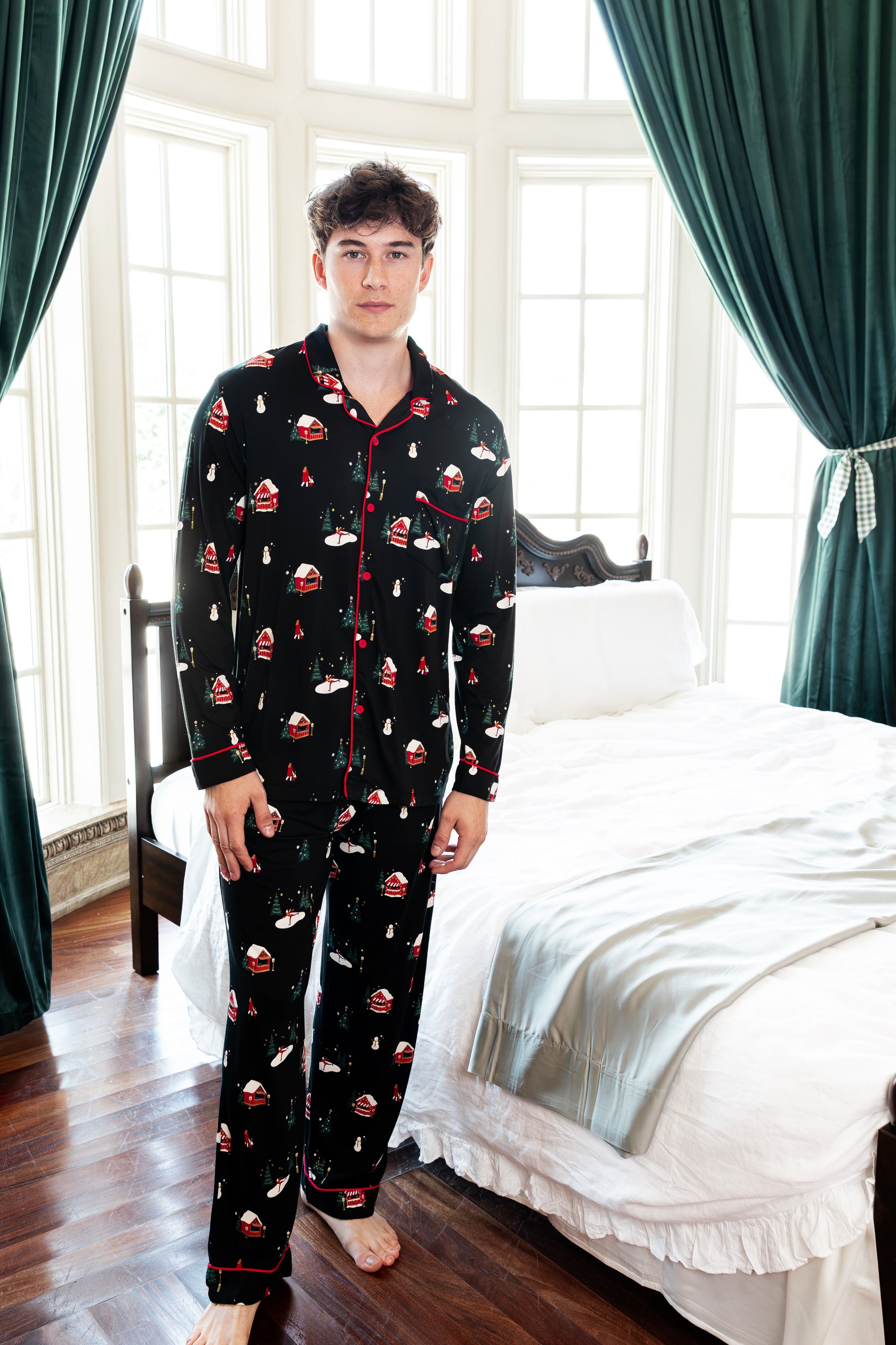 A man wearing a black long-sleeve pajama set with a red and white Christmas-themed print, standing in a bedroom with a bed and green curtains in the background.