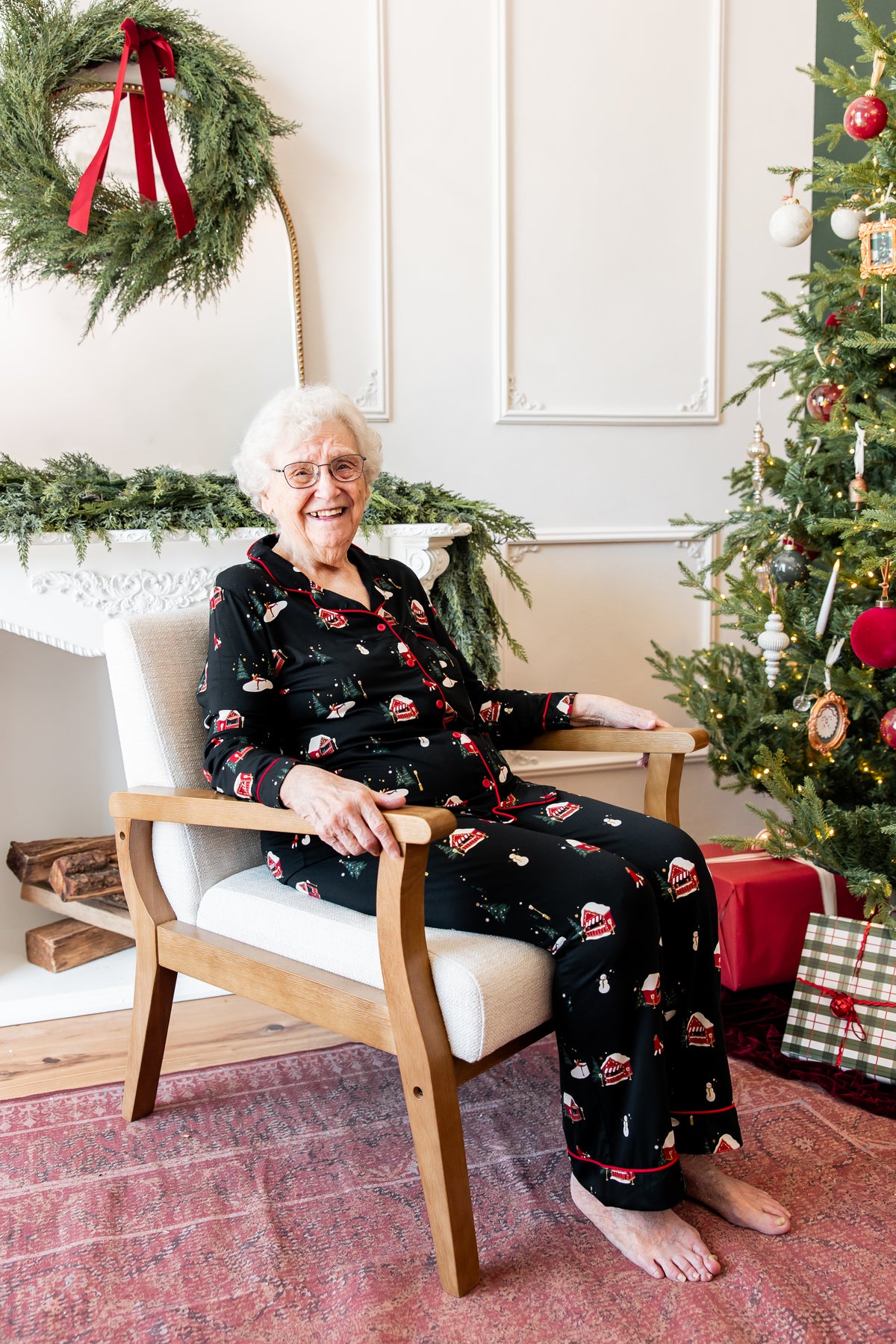 An elderly woman with white hair and glasses sits barefoot in a wooden armchair wearing a black Christmas-themed pajama set with red and white print. The setting is a decorated holiday room with a Christmas tree, wreath, and wrapped gifts.