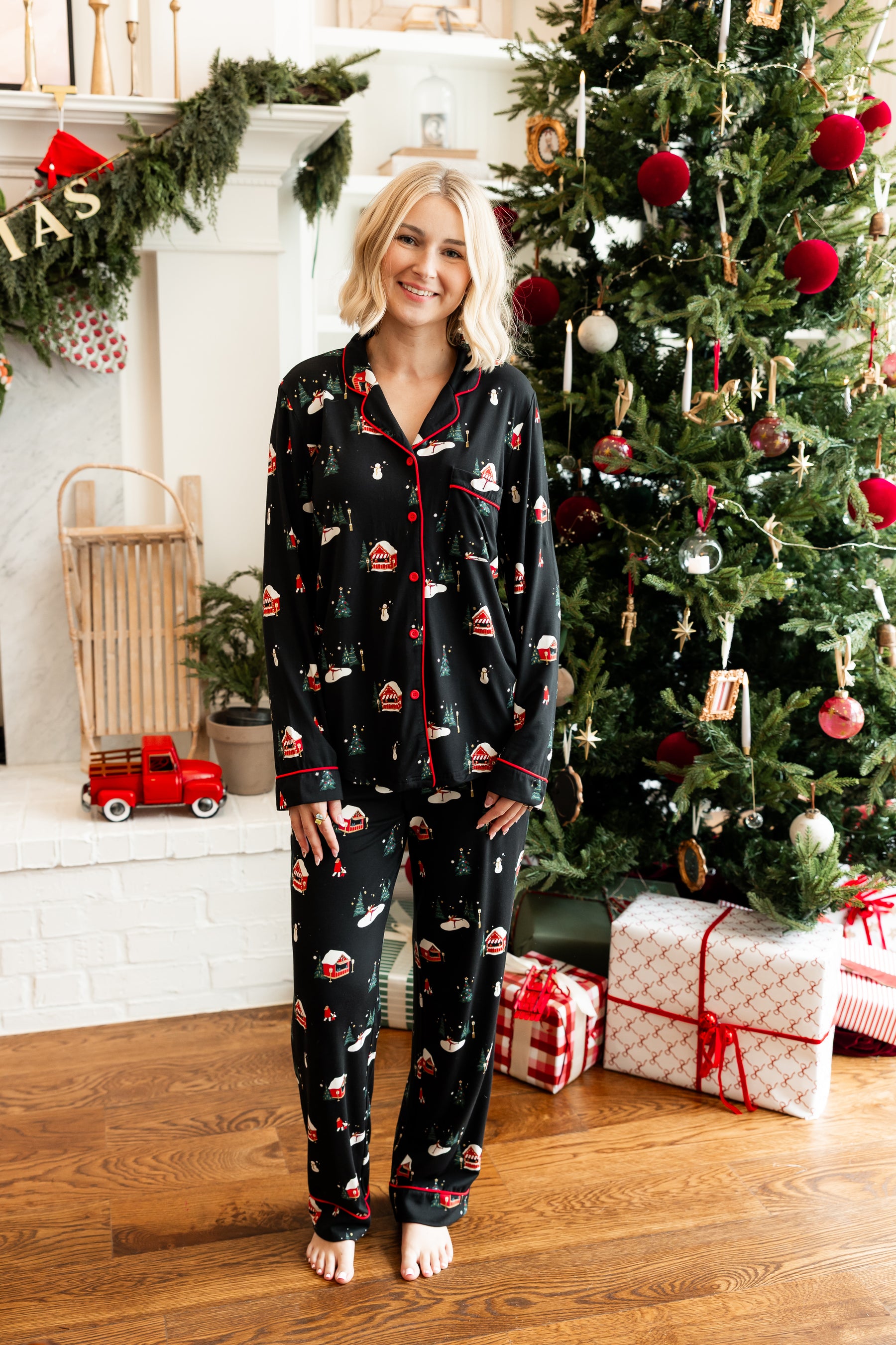 A woman stands barefoot in a living room wearing a black pajama set with a Christmas print, positioned in front of a decorated Christmas tree and fireplace.
