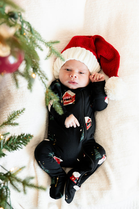 A baby lying on a soft cream blanket, wearing a red and white Santa hat and a black Christmas-themed footed onesie with holiday graphics. Christmas tree branches and ornaments are visible on the left side of the frame.