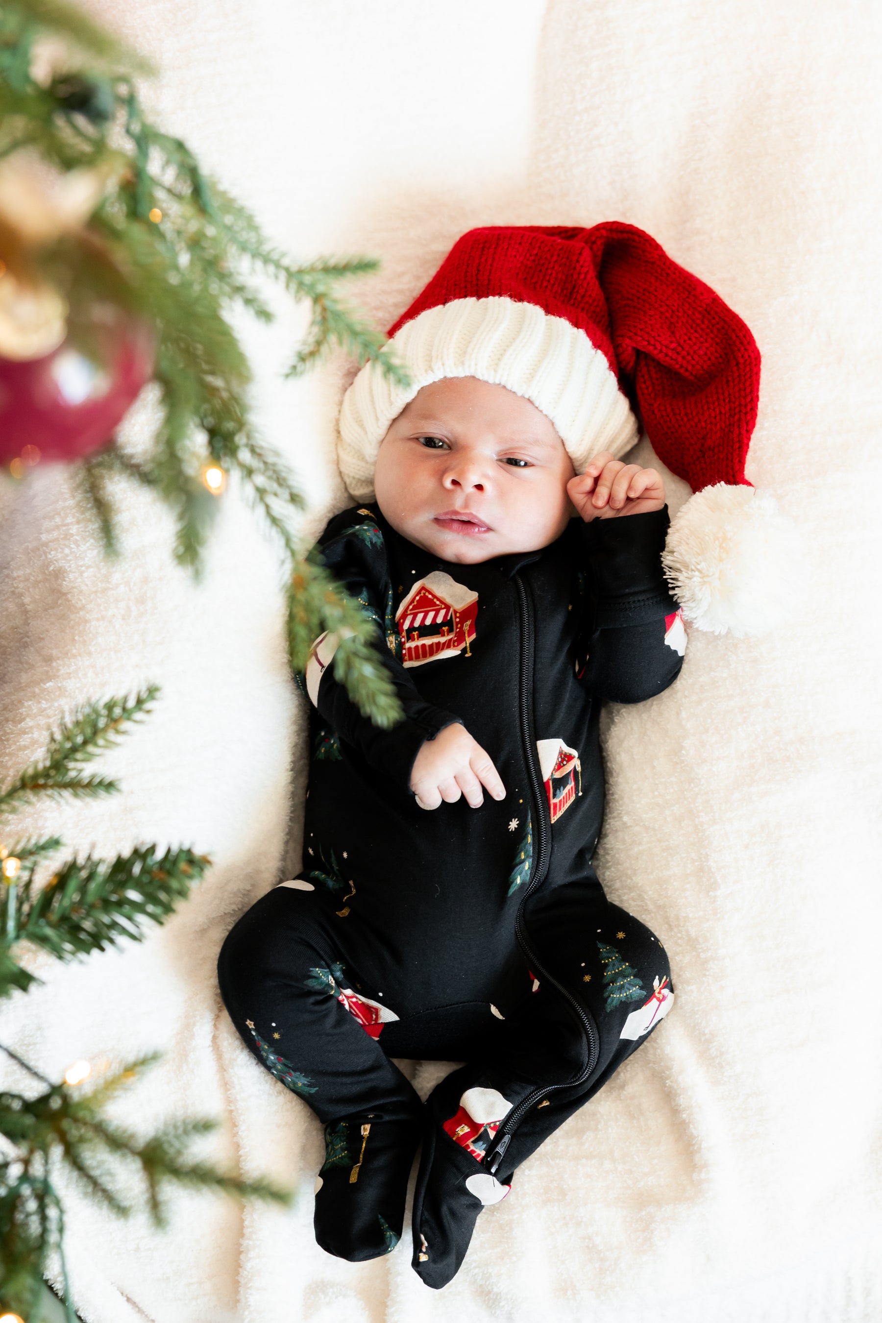 A baby lying on a soft cream blanket, wearing a red and white Santa hat and a black Christmas-themed footed onesie with holiday graphics. Christmas tree branches and ornaments are visible on the left side of the frame.