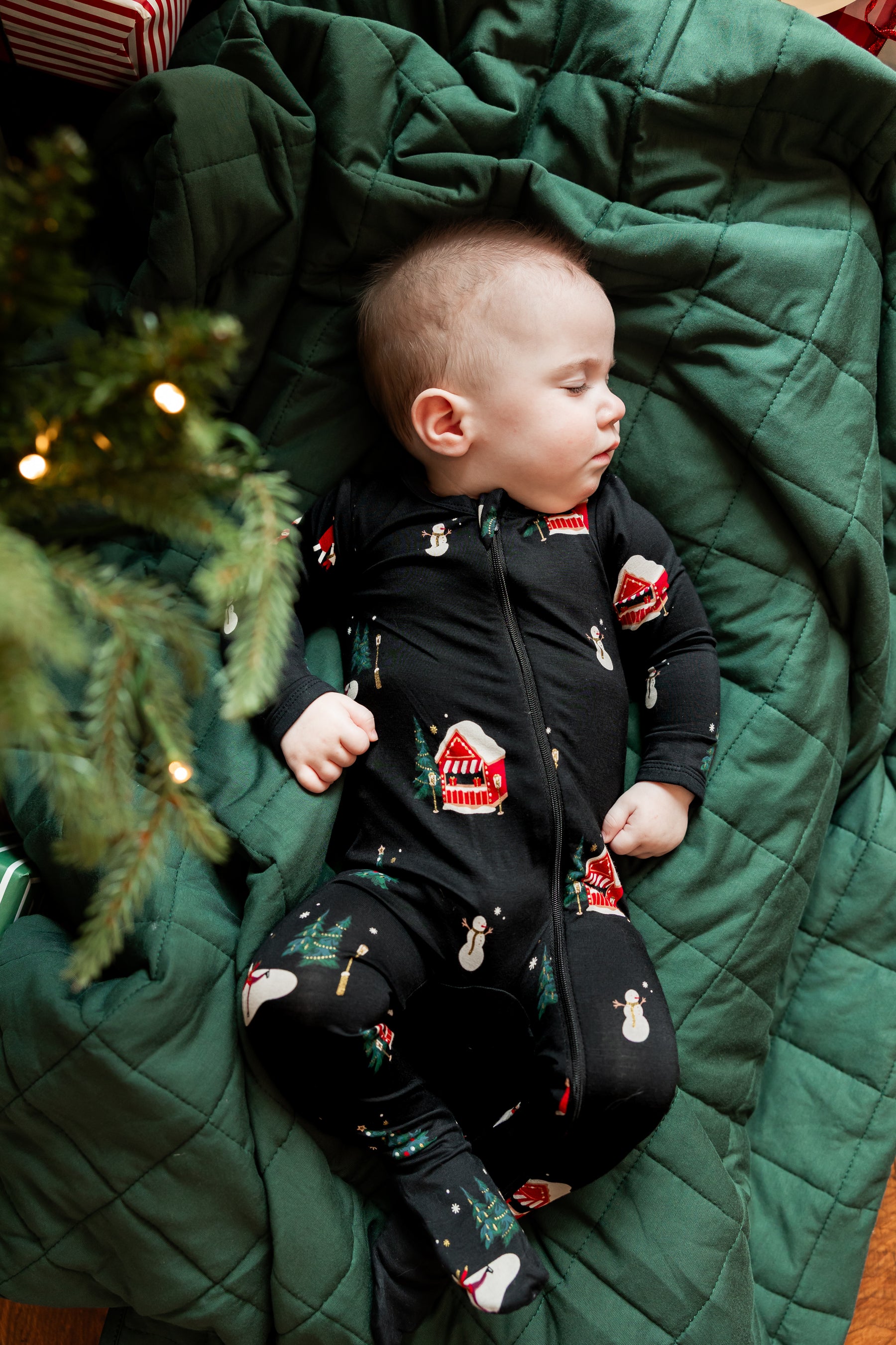 A baby wearing a black zip-up footed onesie with a Christmas print, lying on a dark green quilted blanket next to a Christmas tree.