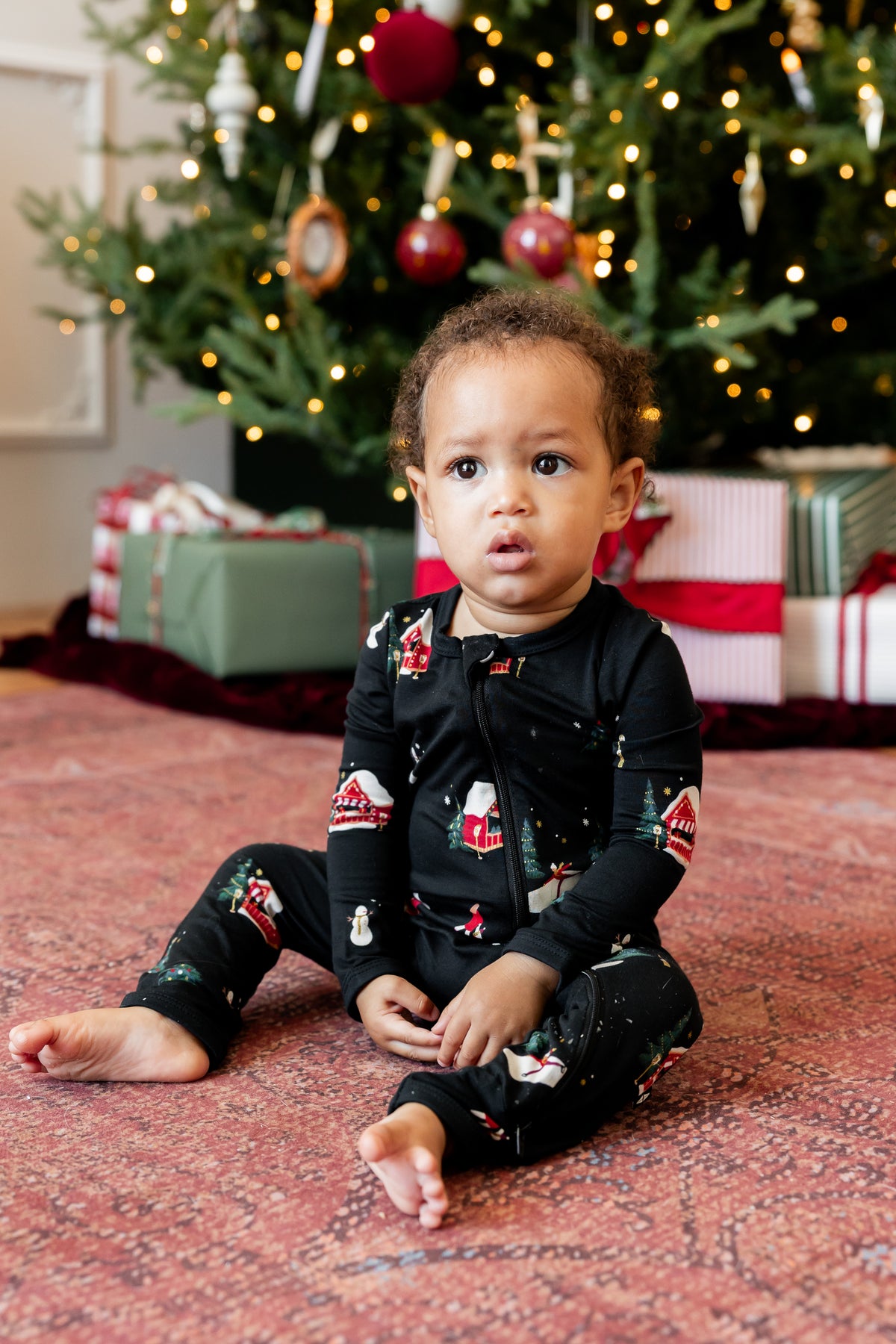A toddler sitting on a patterned red rug in front of a decorated Christmas tree, wearing a black long-sleeve pajama set with a Santa Claus and snowman print.