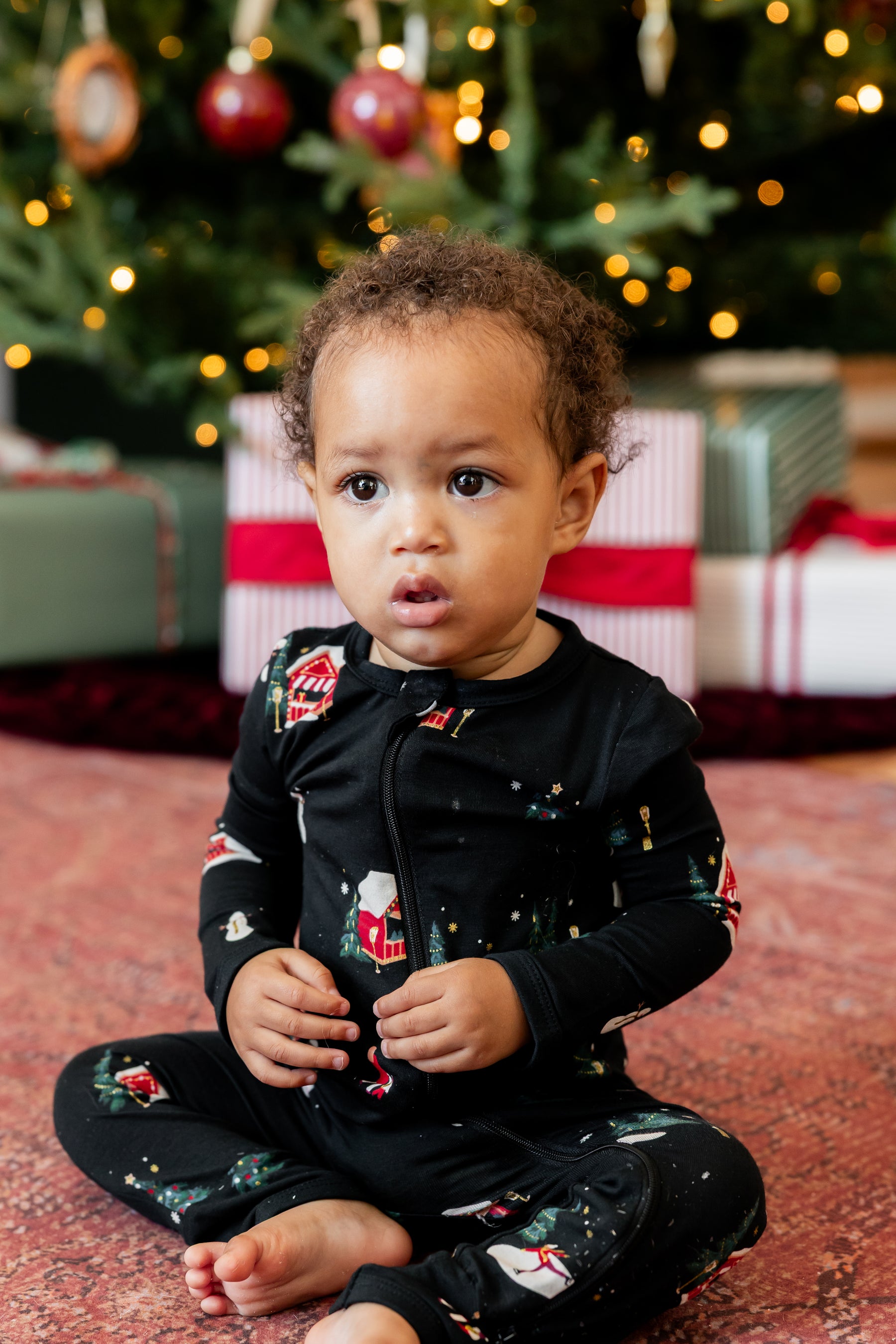 A toddler sitting on a patterned rug in front of a decorated Christmas tree and wrapped gifts, wearing a black long-sleeve pajama set with a festive holiday print.
