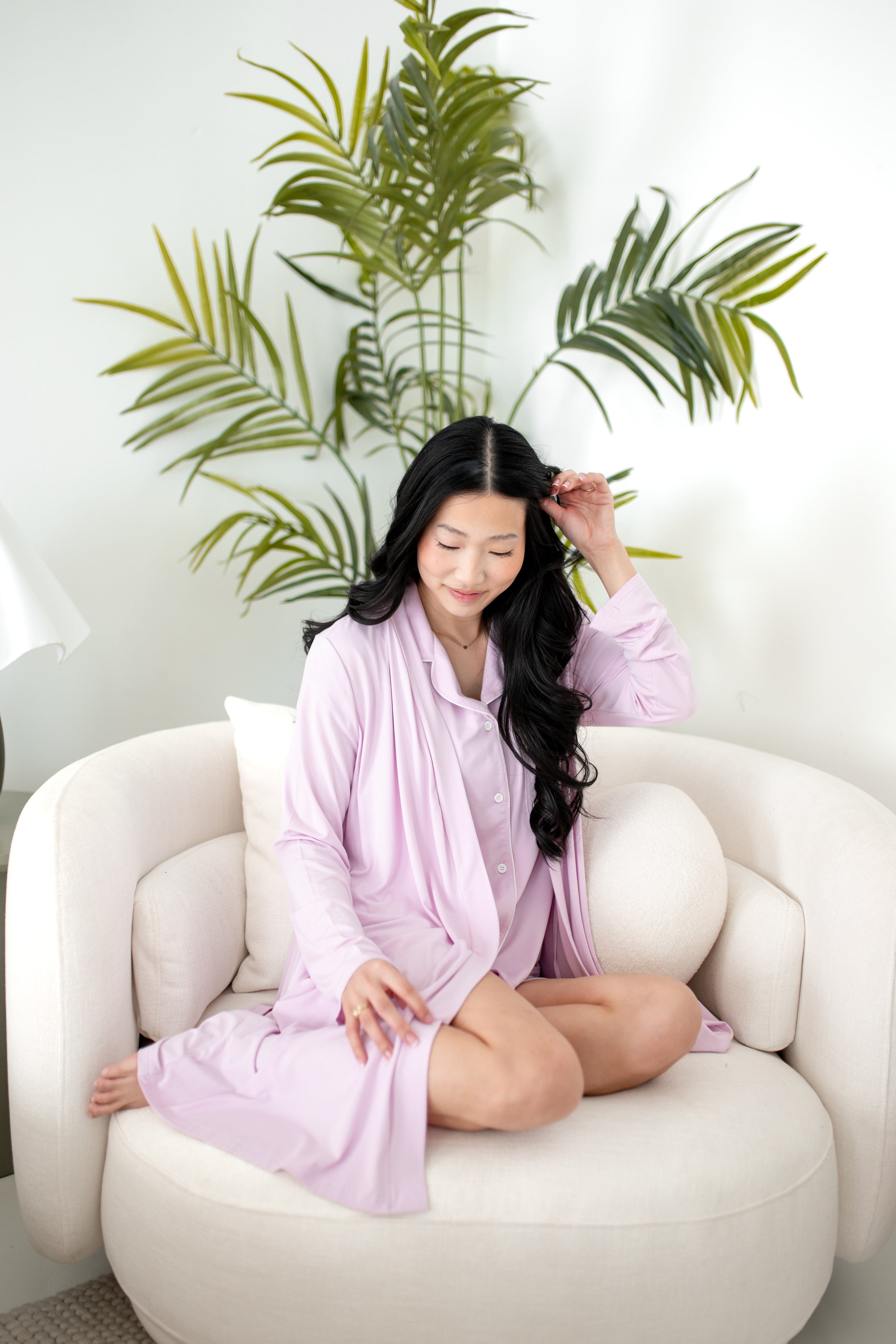 Woman wearing lounge robe and short sleeve PJ set in a light purple color sitting on a chair with plant in the background