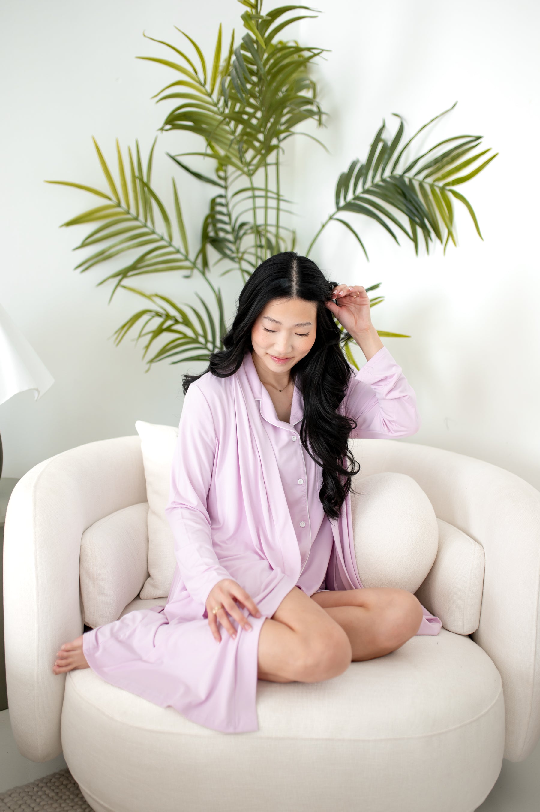 Woman wearing lounge robe and short sleeve PJ set in a light purple color sitting on a chair with plant in the background