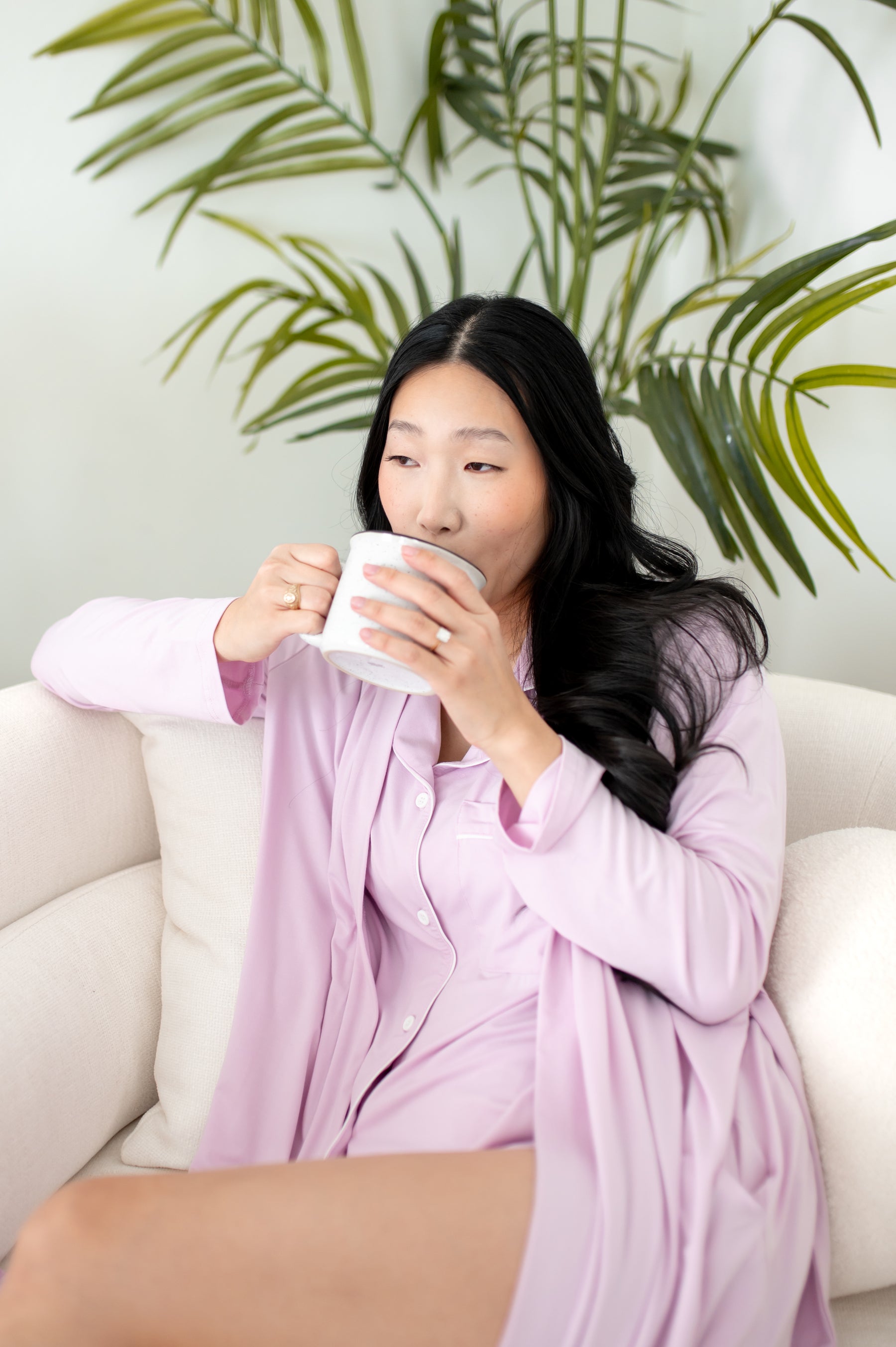 Woman in purple pajamas sitting on a white couch holding a mug with green plants in the background.