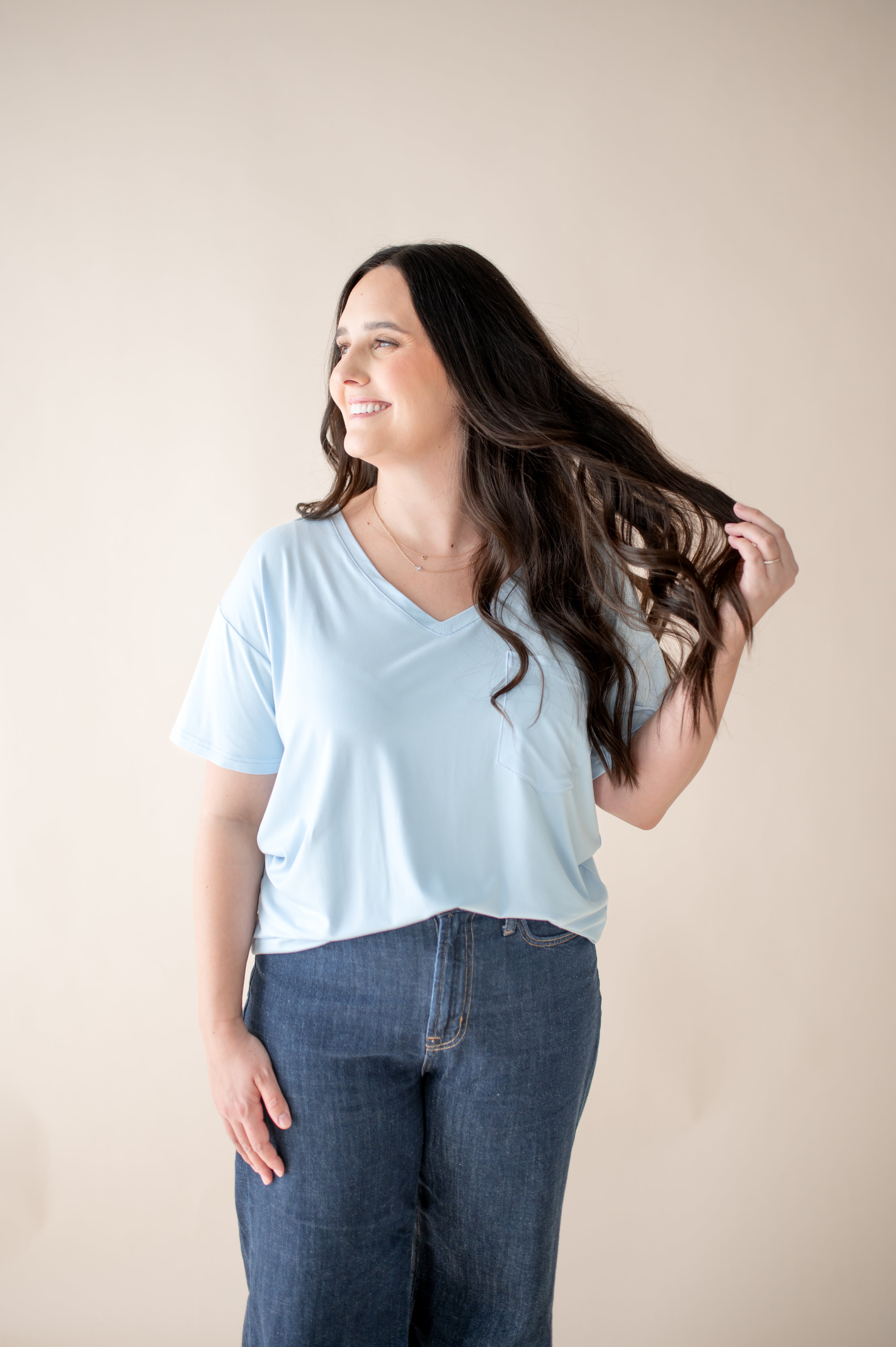 Female standing in front of a light neutral background with her hand in her hair wearing the Women's Relaxed Fit V-Neck in Breeze paired with medium wash jeans