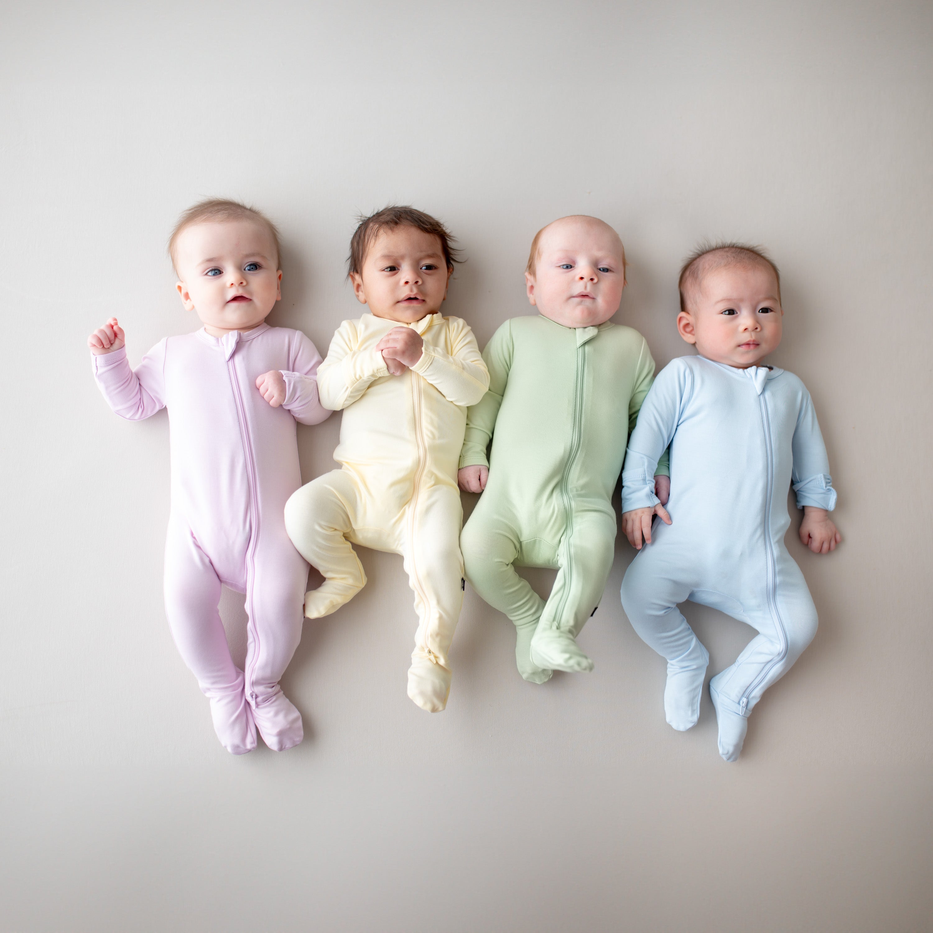 Four babies wearing pastel-colored footies on a neutral background