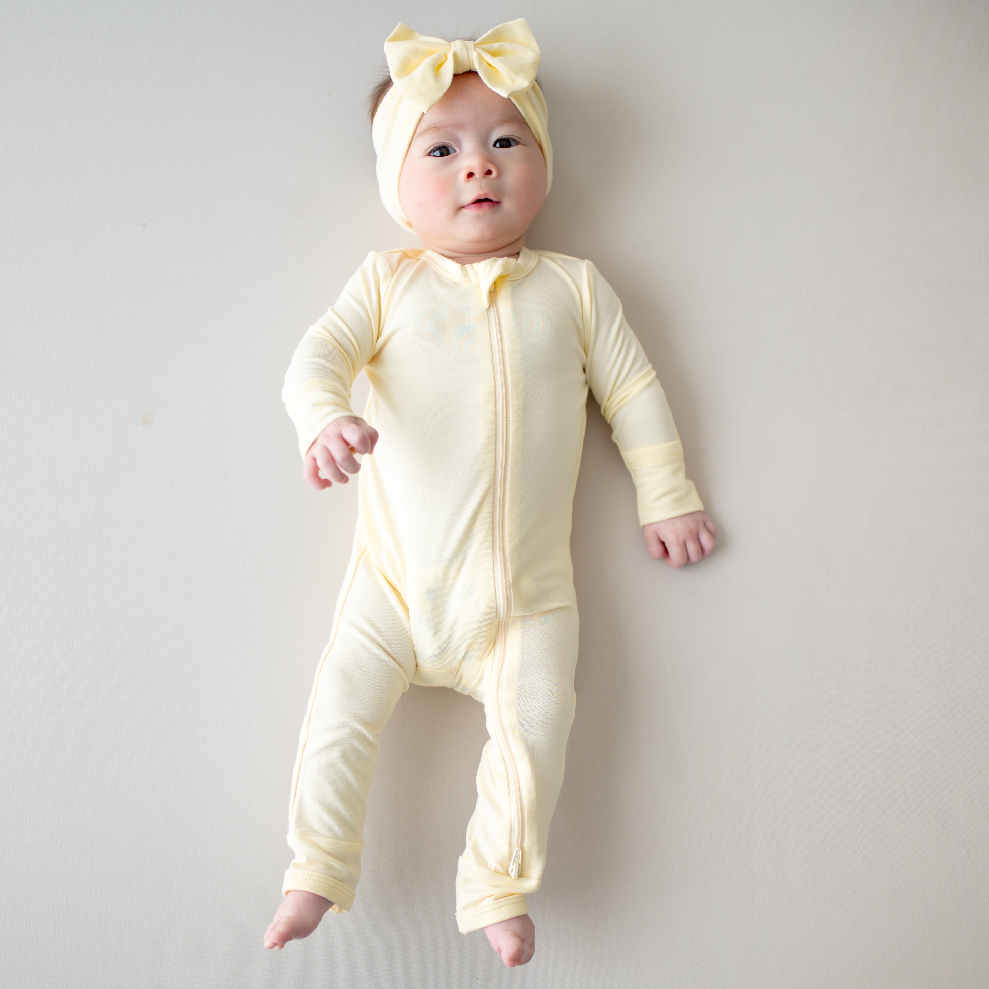 Baby wearing a yellow zippered romper and headband on a plain background
