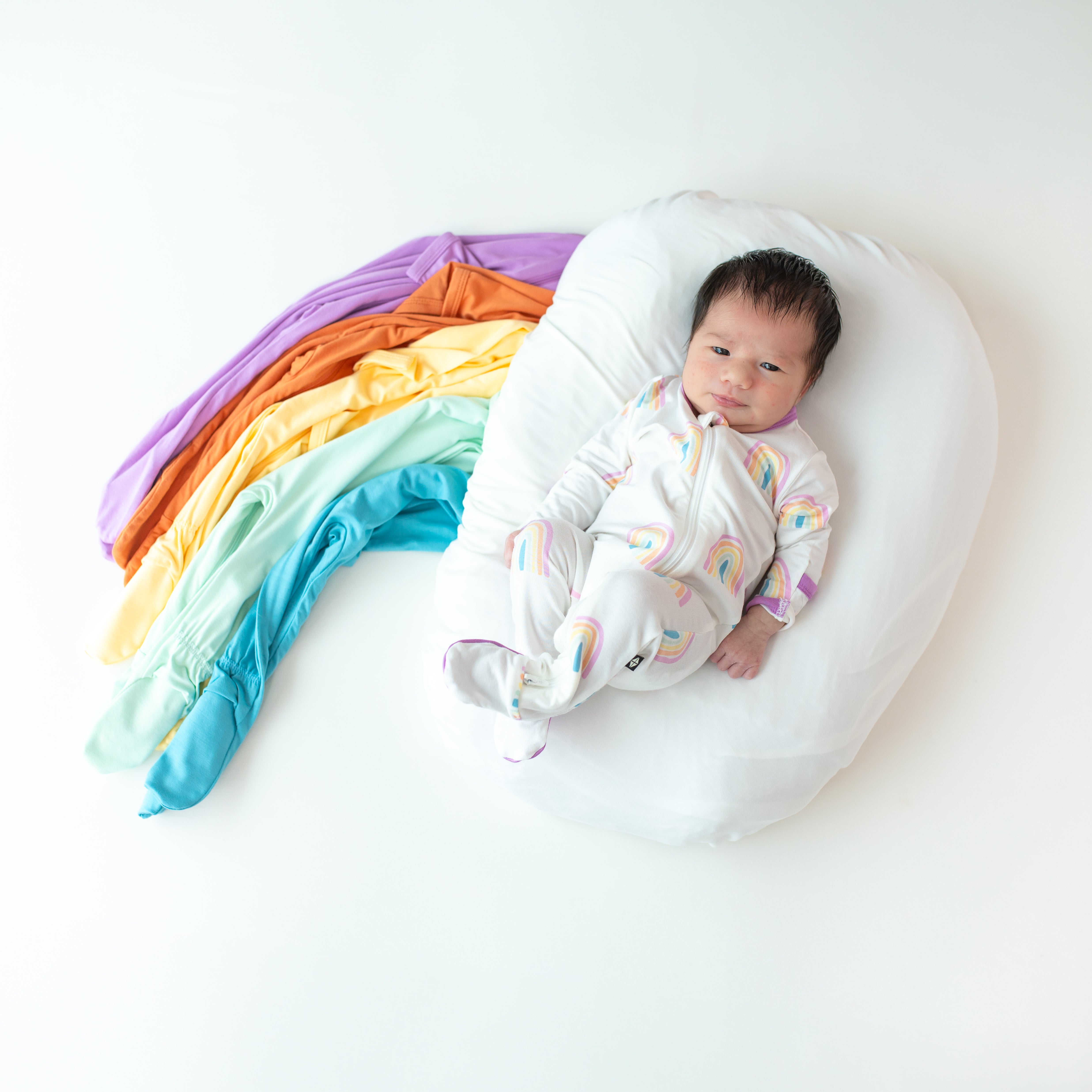 Baby in Zippered Footie in Poi Rainbow next to a rainbow made of footies