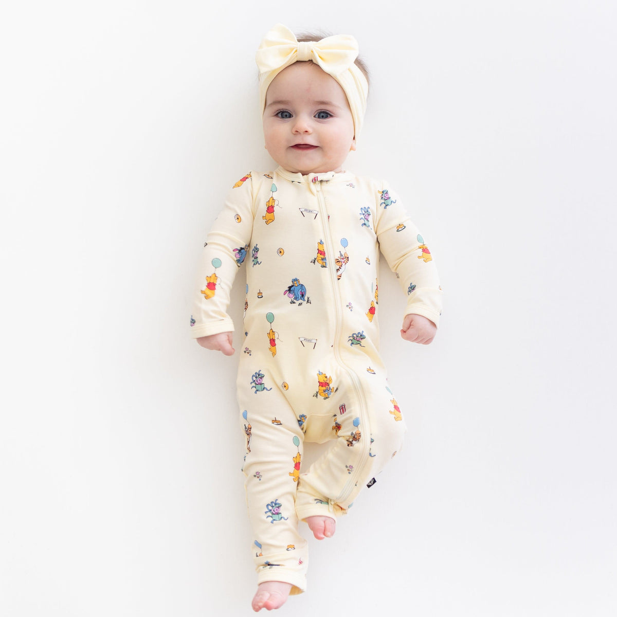 Young toddler laying on a light neutral surface wearing the Zippered Romper in Winnie the Pooh Celebration with a light yellow bow
