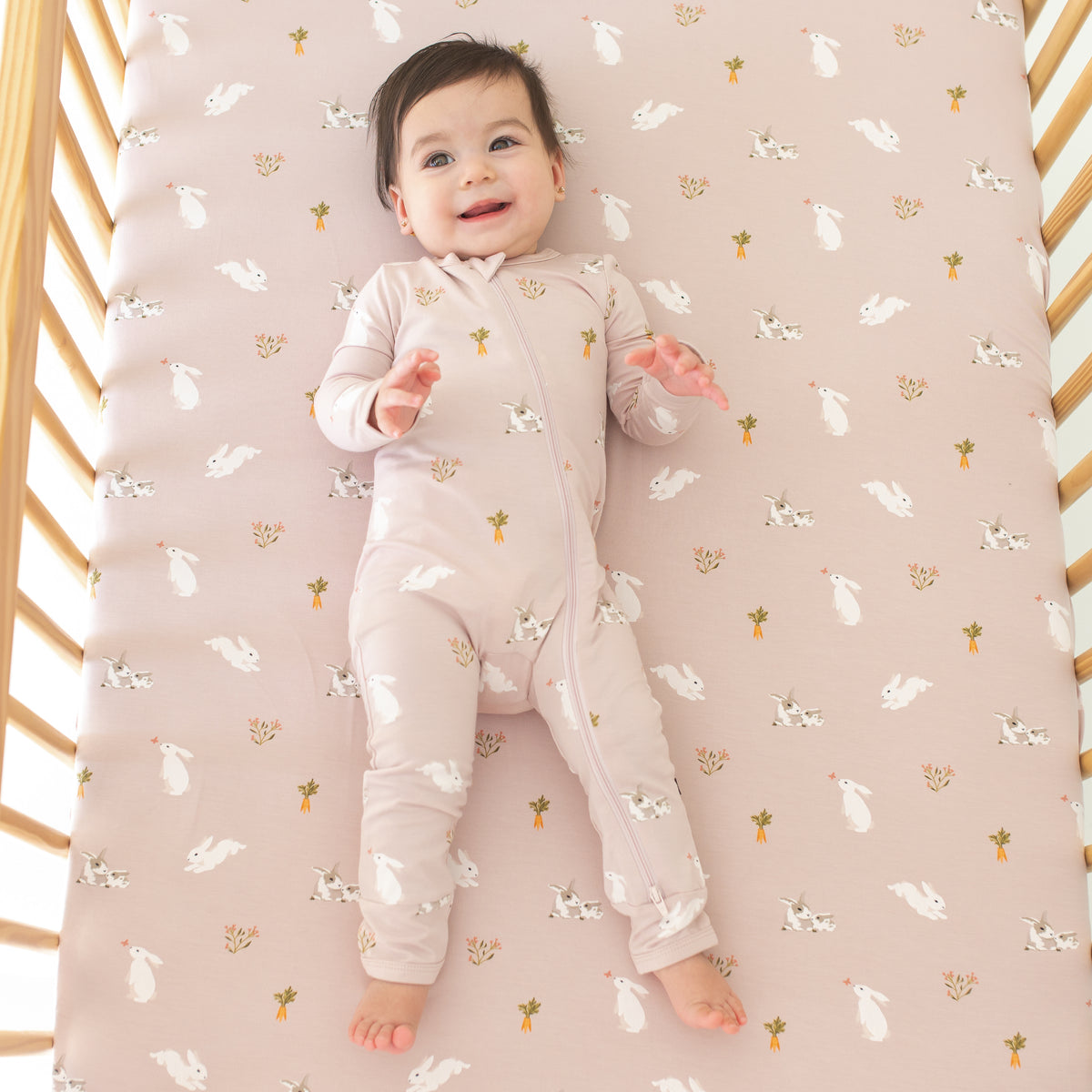 Baby wearing Zippered Romper in Blush Rabbit laying in crib with Blush Rabbit crib sheet