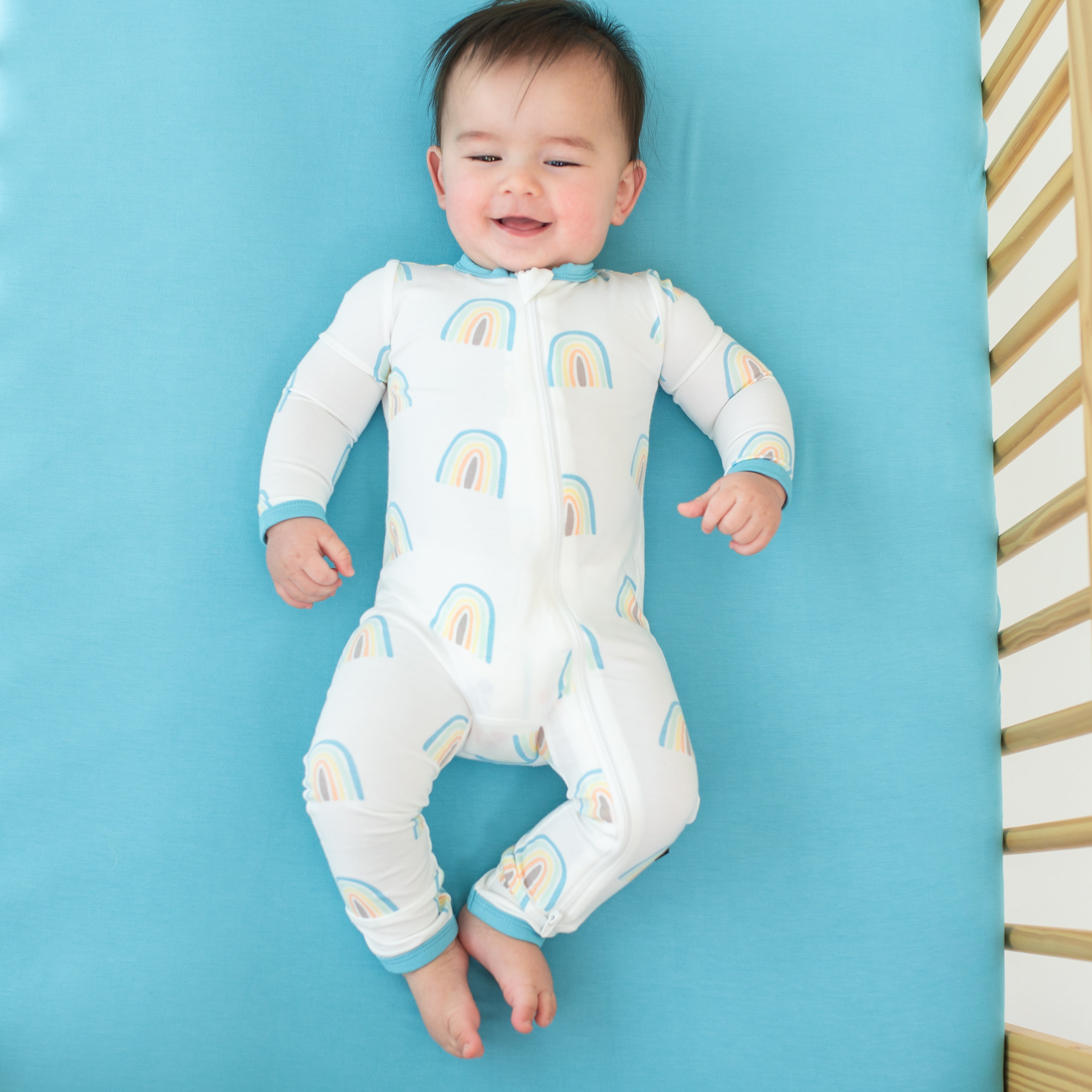 Baby smiling wearing Zippered Romper in Makai Rainbow while in a crib with makai crib sheet