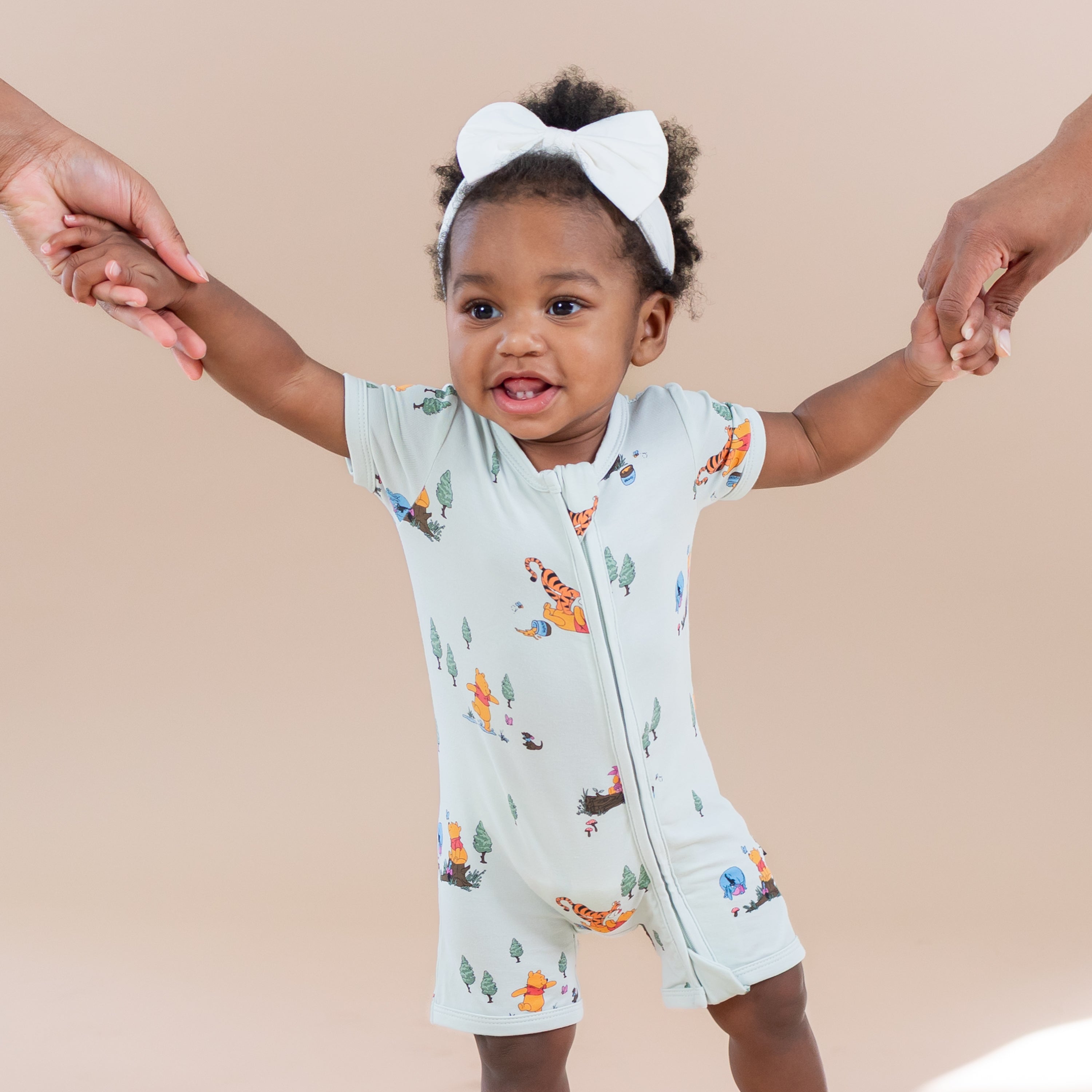Toddler girl standing holding her moms hands wearing the Zipper Shortall Romper in Winnie the Pooh Hundred Acre Wood with a white bow