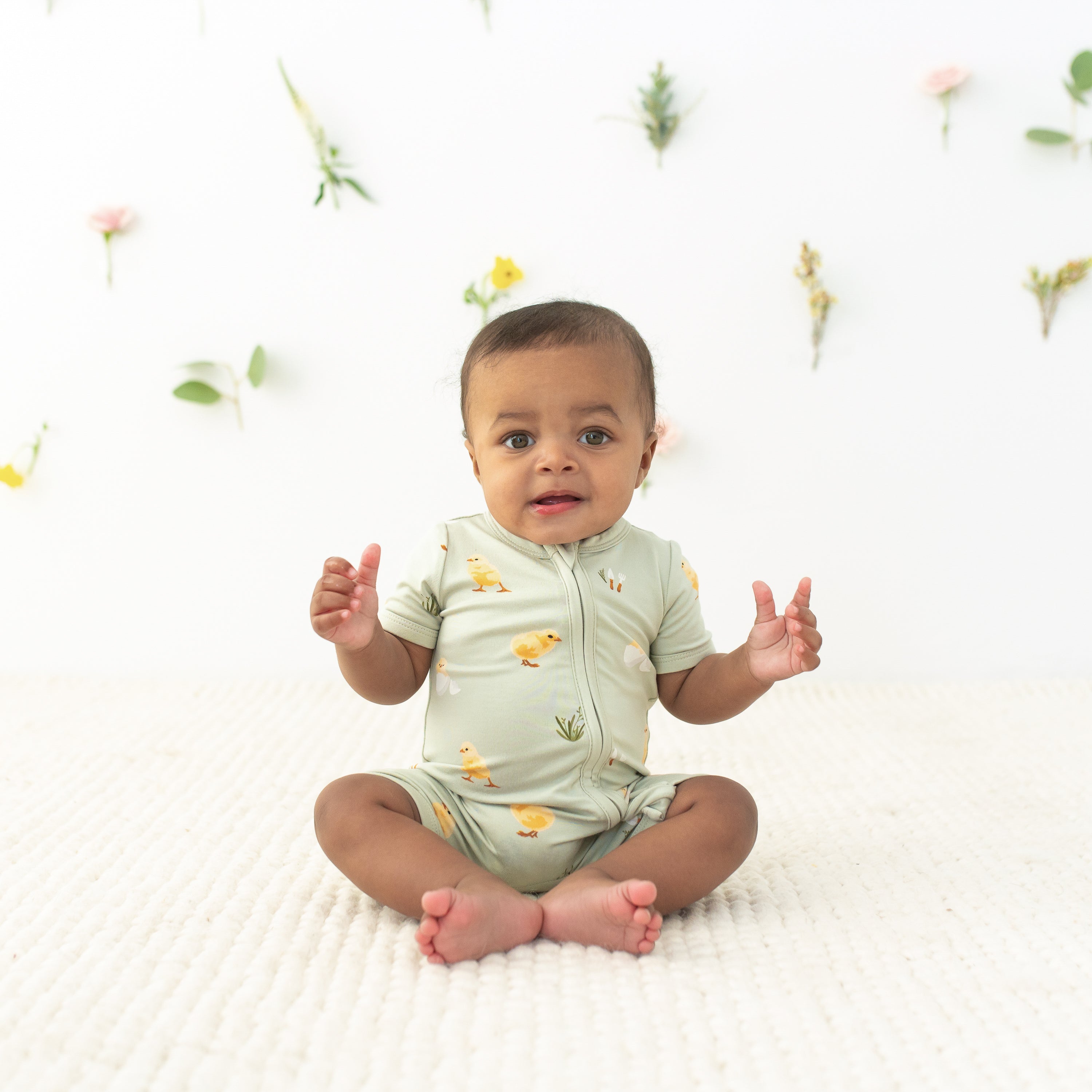 Baby sitting wearing Zipper Shortall Romper in Aloe Chick with floral background