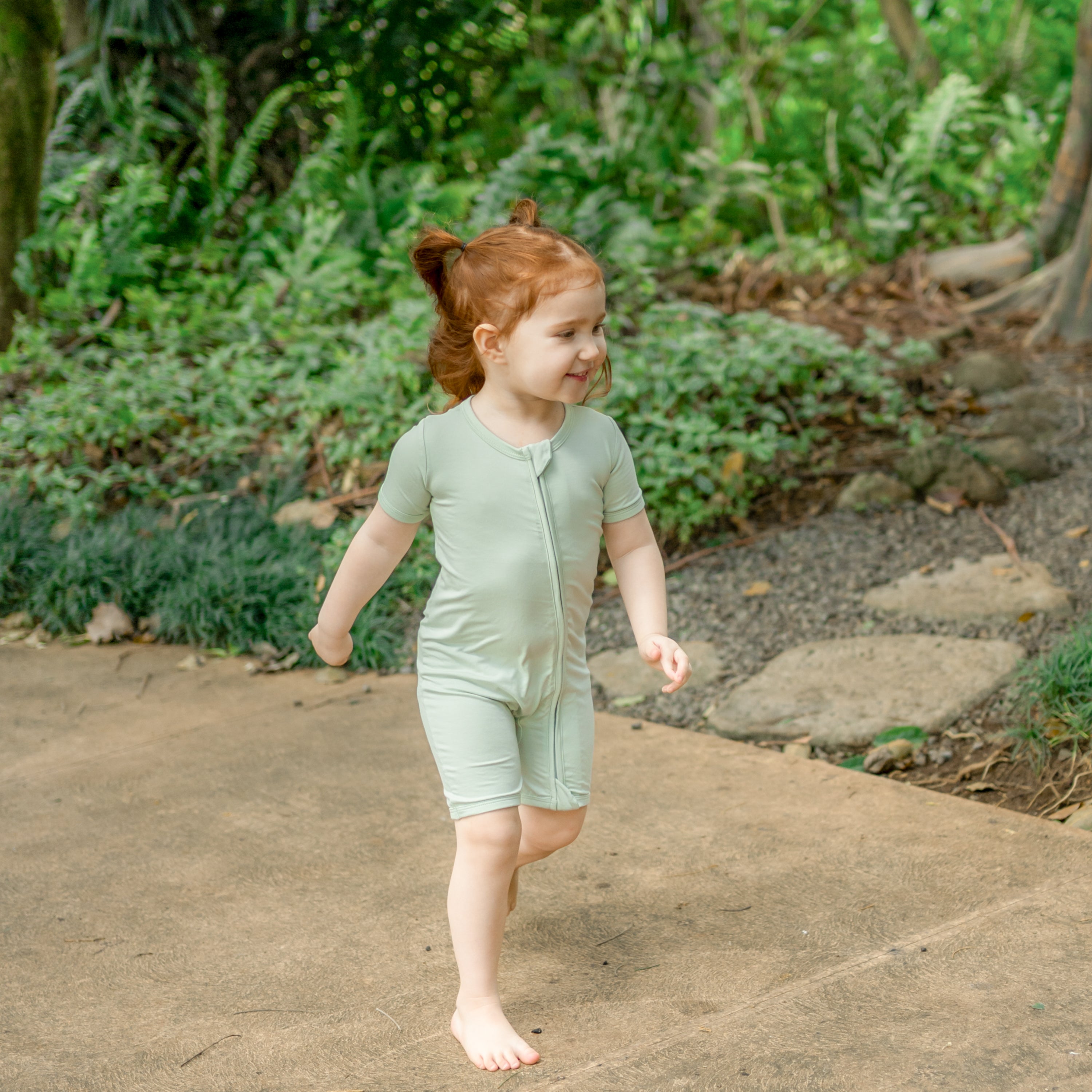 Young girl walking on a path wearing the Zipper Shortall Romper in Basil