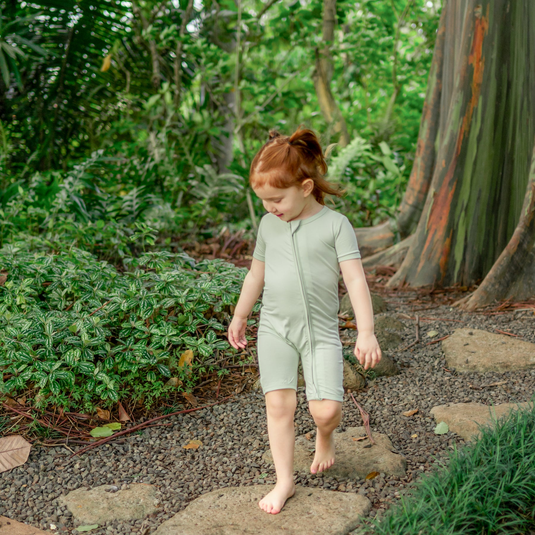 Young girl walking outside on a stone path wearing the Zipper Shortall Romper in Basil