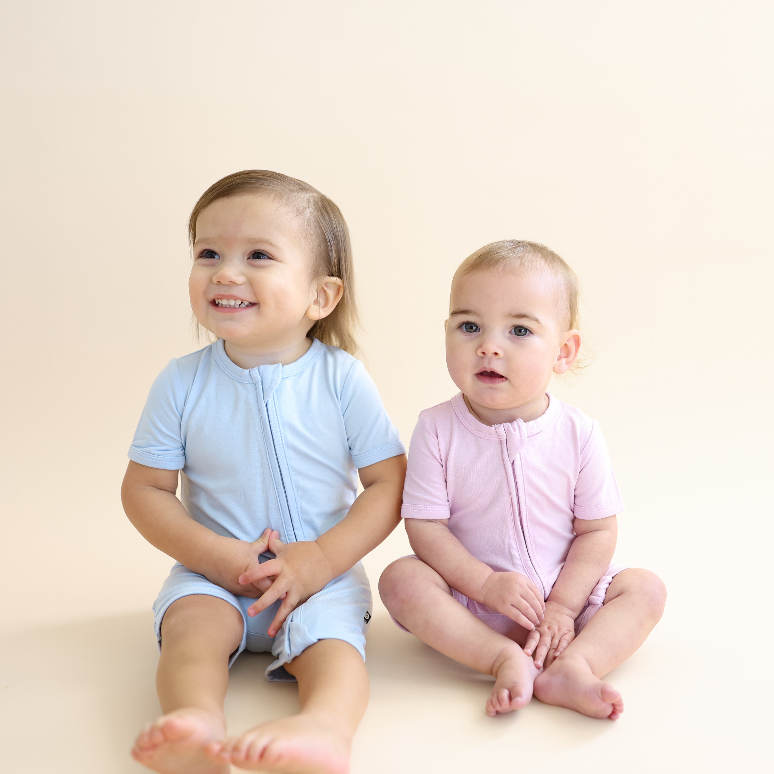 Two young toddlers sitting on the floor wearing the Zipper Shortall Romper in Breeze and Thistle