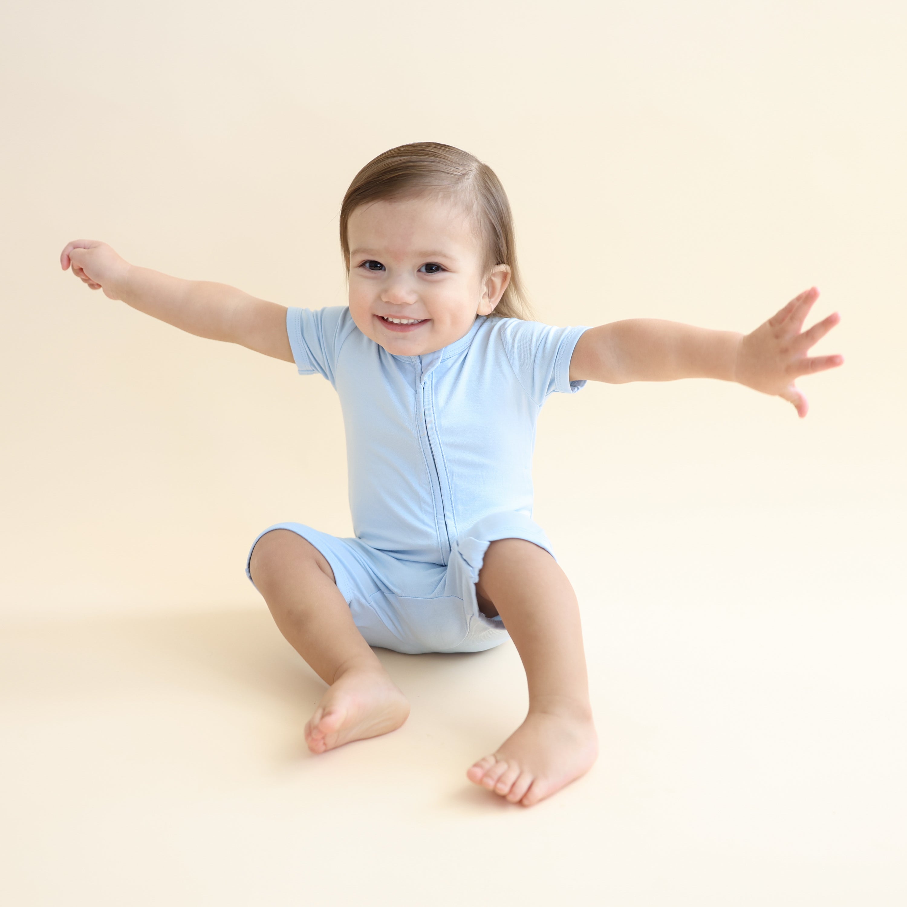 Young boy sitting on the floor with his arms at his side wearing the Zipper Shortall Romper in Breeze