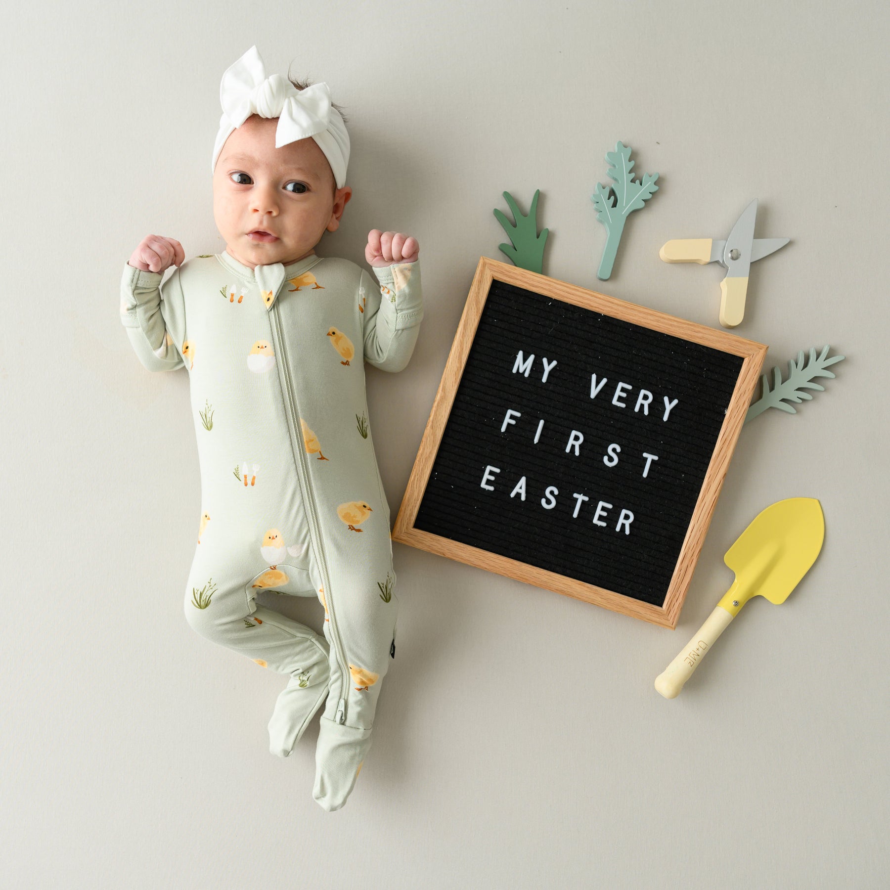 Baby wearing Zippered Footie in Aloe Chick next to a first easter sign