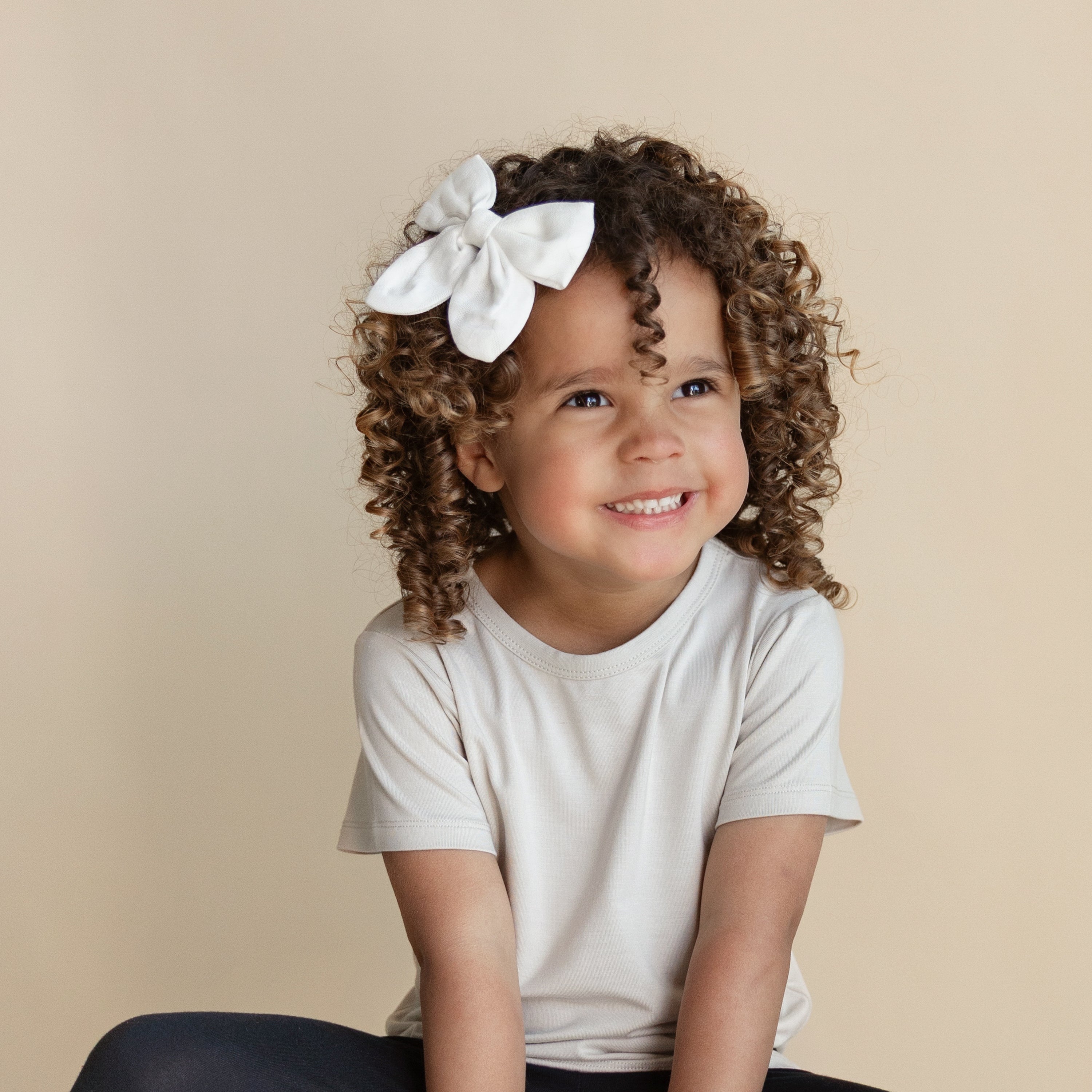 Toddler wearing Kyte Baby's Oat Basic Tee with black leggings and a white hair bow