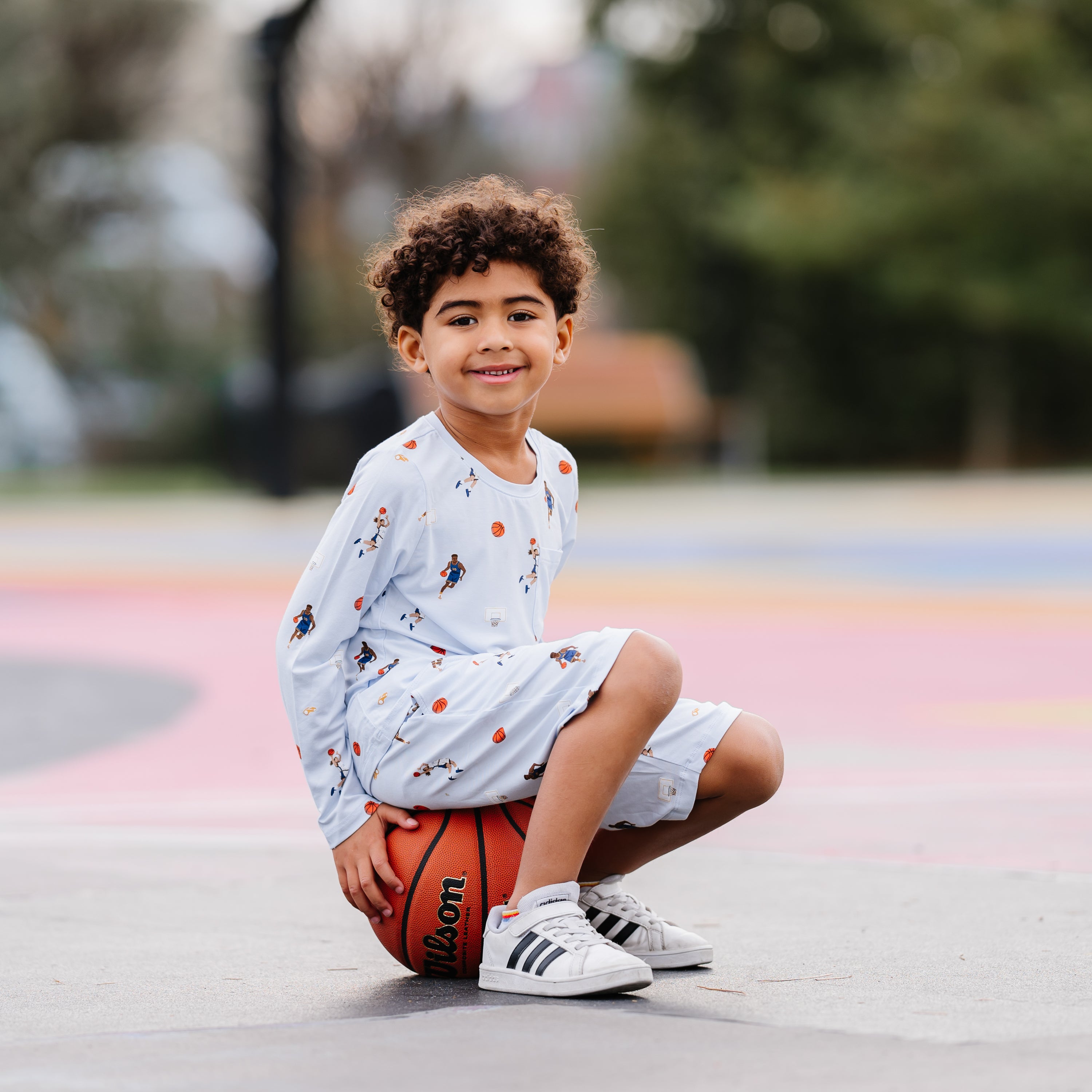 Young boy sitting on a basketball wearing the Long Sleeve Toddler Crew Neck Tee in Basketball with matching drawstring shorts