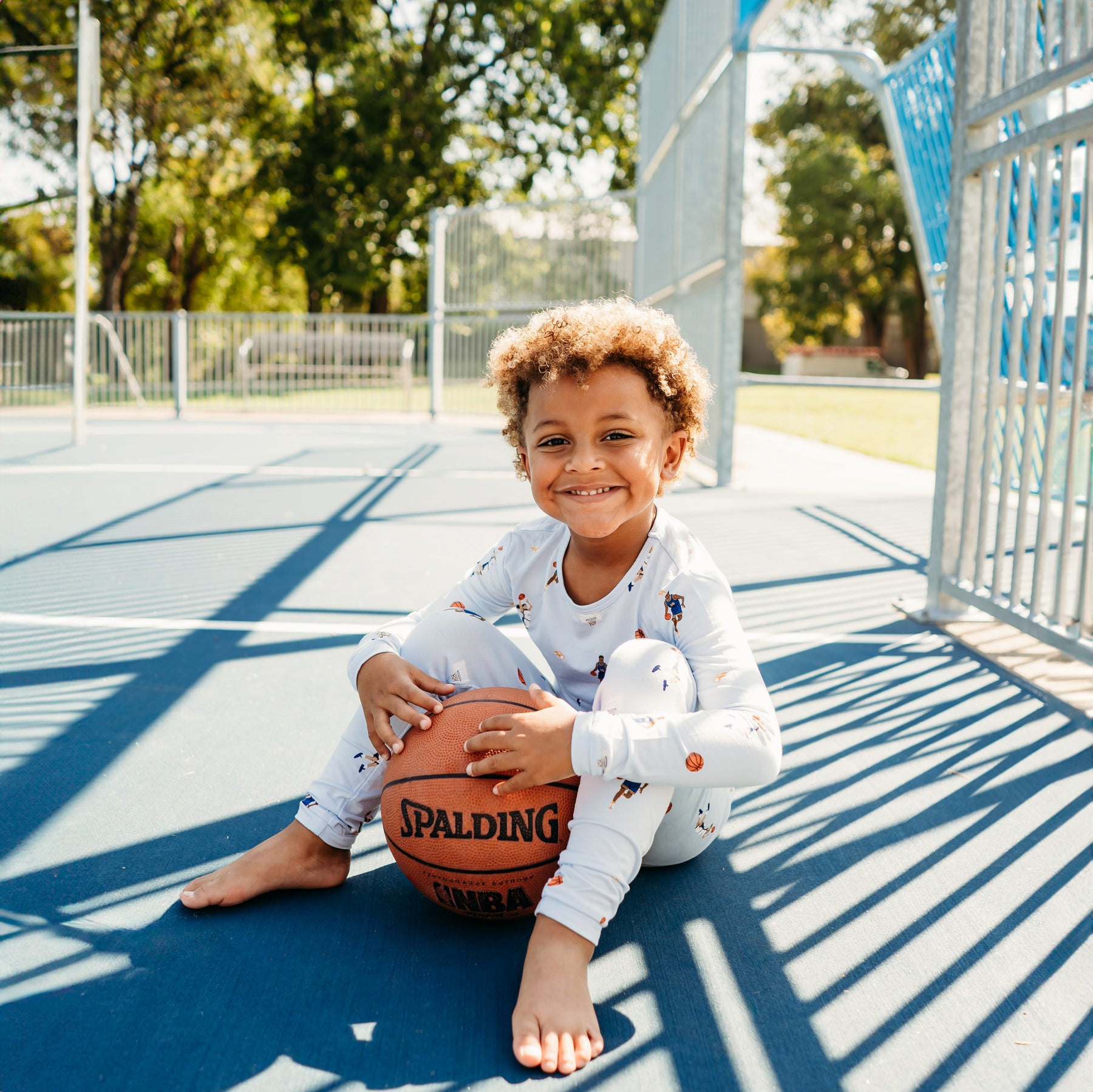 Young smiling boy sitting on a basketball court wearing the Long Sleeve Pajamas in Basketball holding a basketball