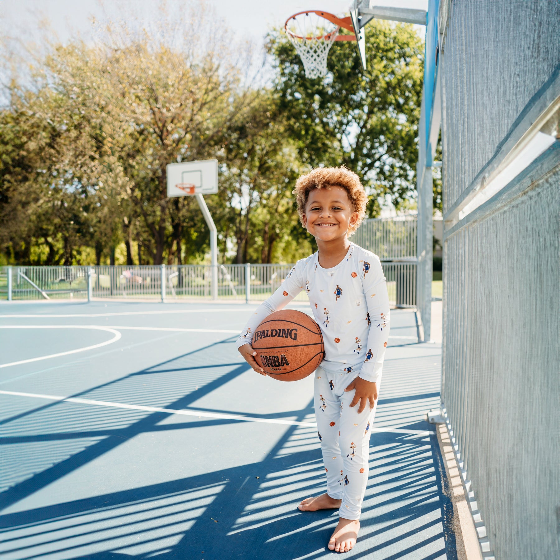 Smiling boy holding a basketball standing on a basketball court wearing the Long Sleeve Pajamas in Basketball