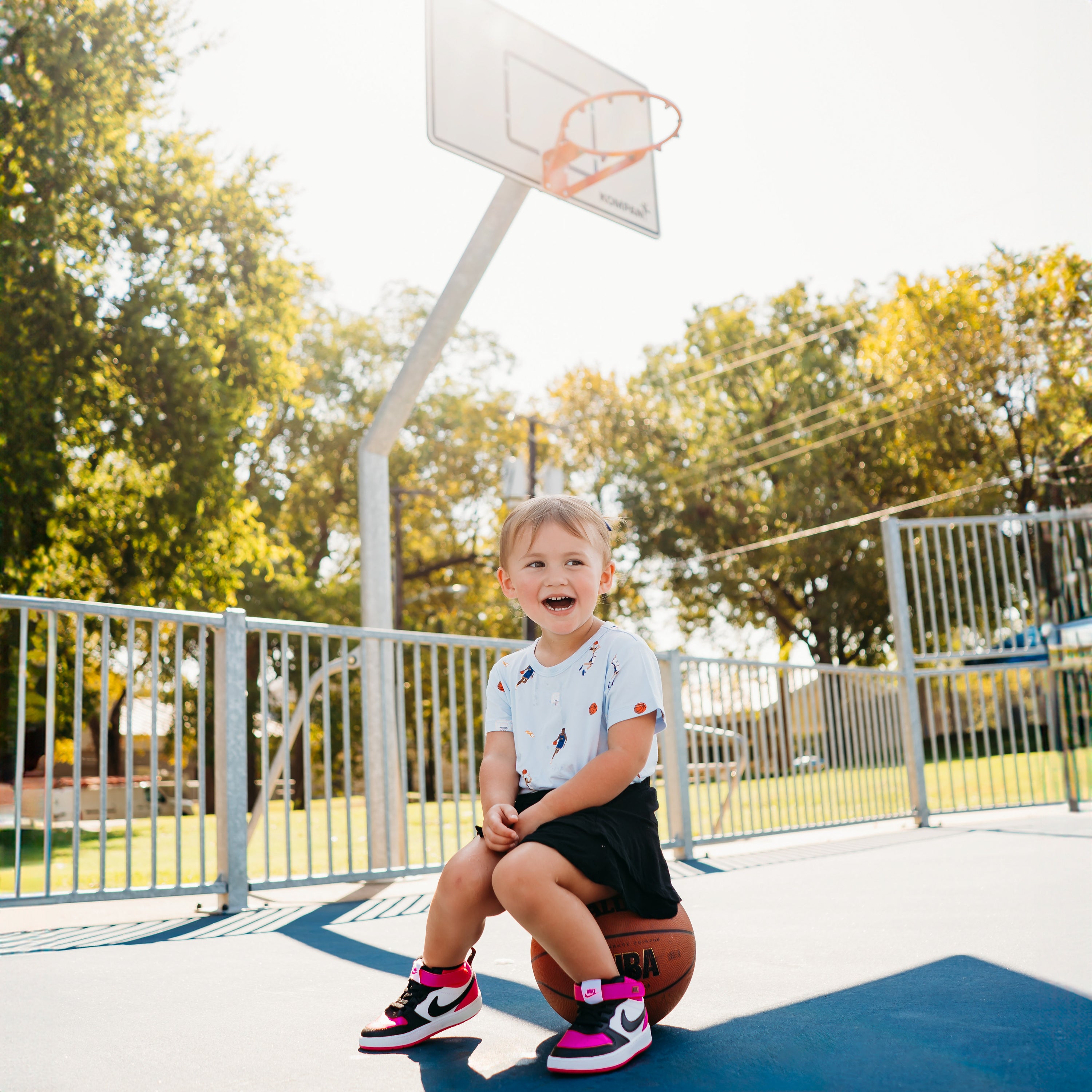 Young girl sitting on a basketball on a basketball court wearing the Toddler Basic Tee in Basketball with a black sikirt
