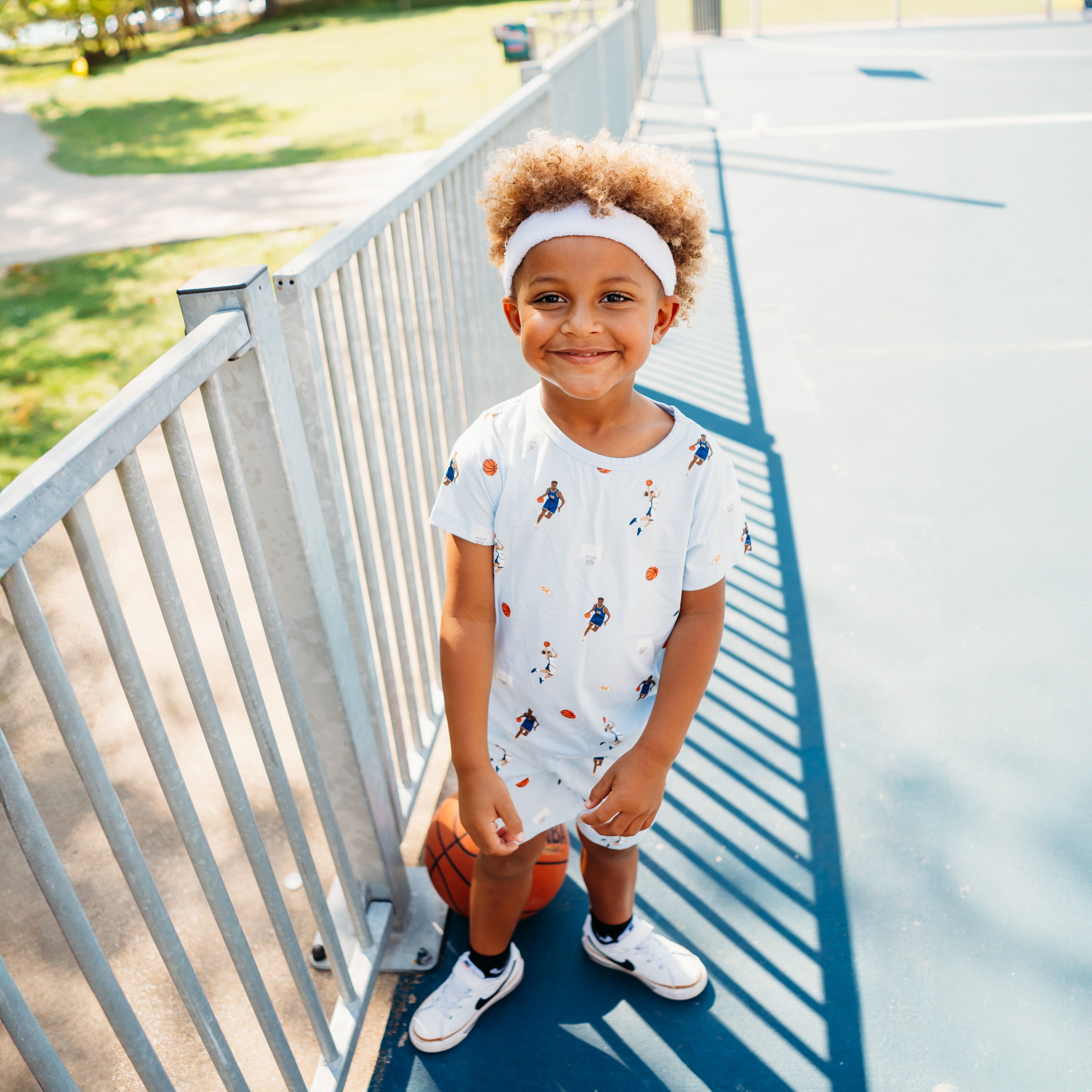 Young boy standing alongside a fence on a basketball court wearing the Toddler Basic Tee in Basketball with a white headband