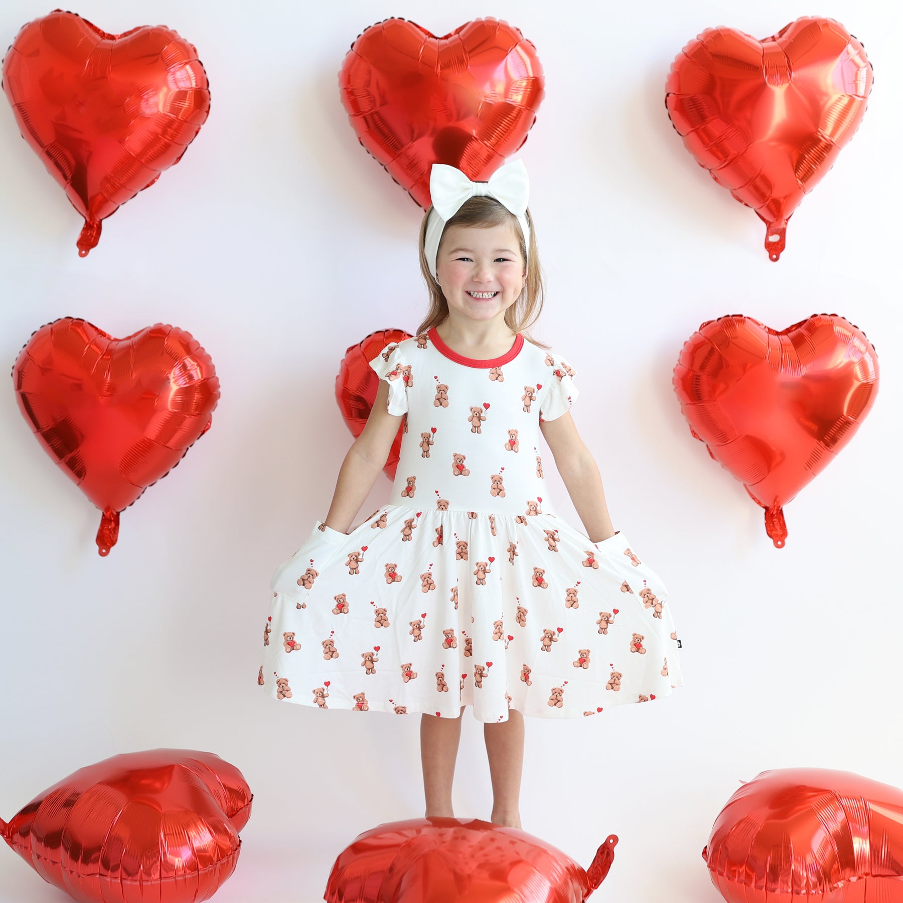 Young girl standing with her hands in the pockets of the Pocket Dress in Bear Hearts surrounded by red heart helium balloons