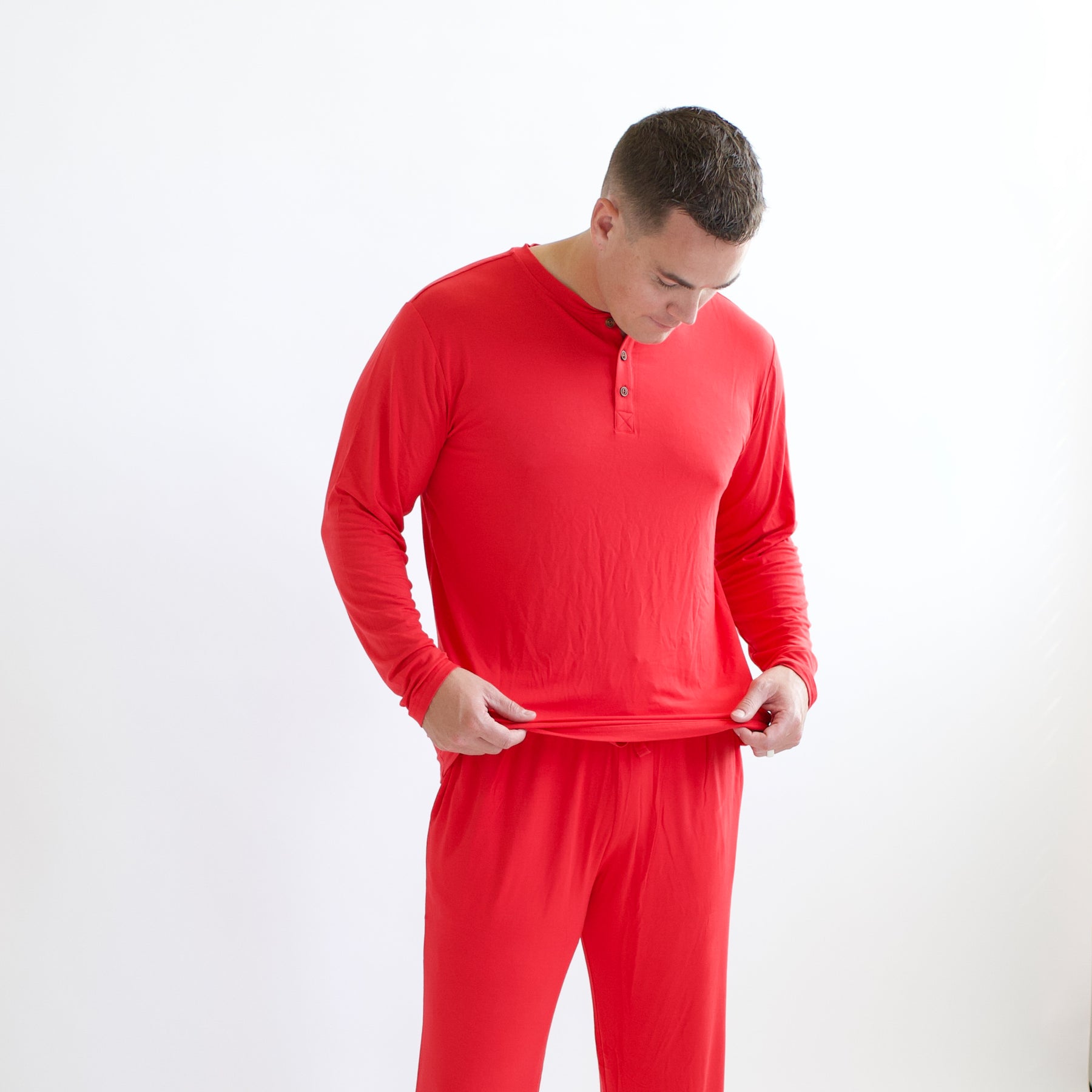Male model  holding the bottom hem of the Men's Long Sleeve Henley Top in Cardinal in his hands standing in front of a white background