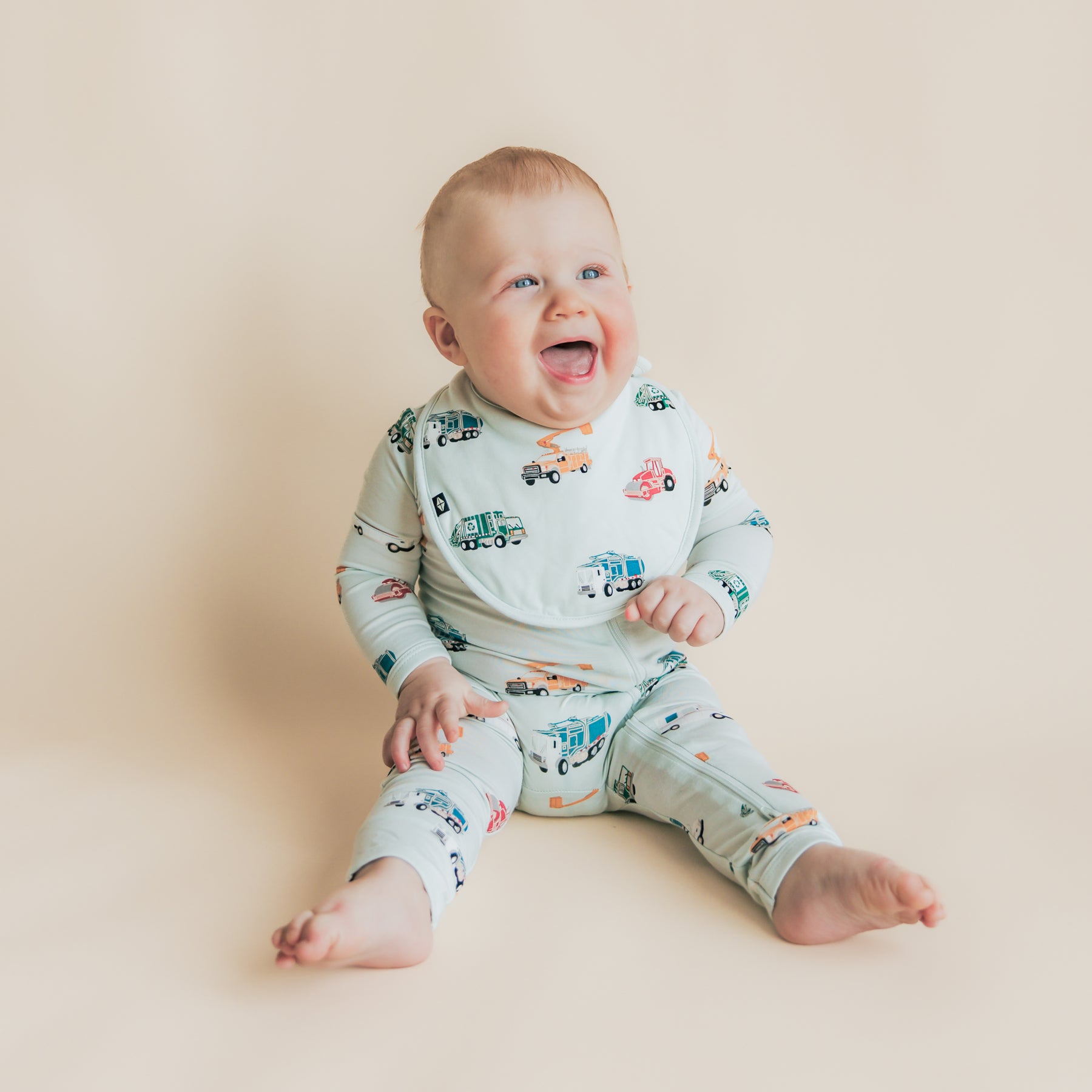Smiling toddler sitting on the floor wearing the Cushy Bib in City Vehicles with matching zippered romper