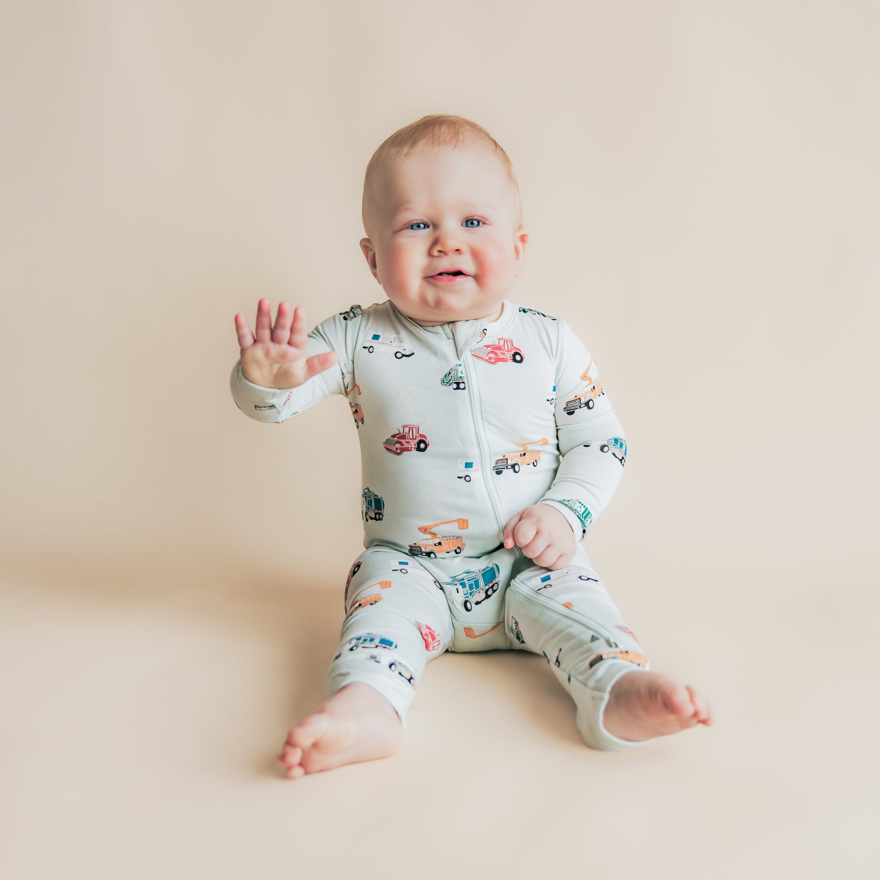 Young toddler sitting on the floor waving wearing the Zippered Romper in City Vehicles