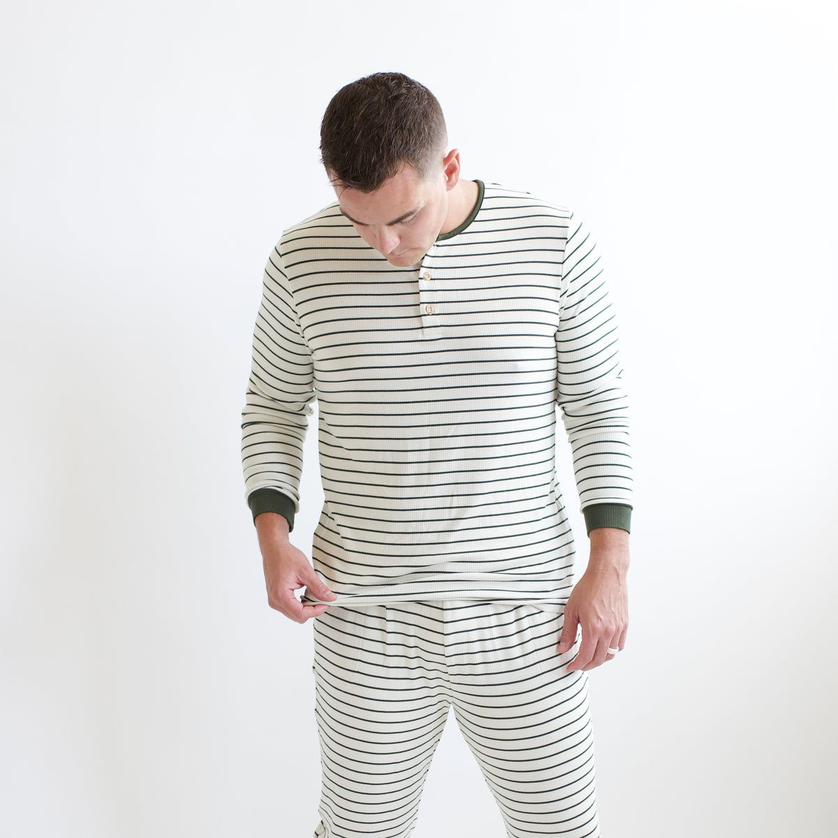 Male model holding the hem of the shirt of the Men's Ribbed Henley Set in Fir Stripe in front of a white background