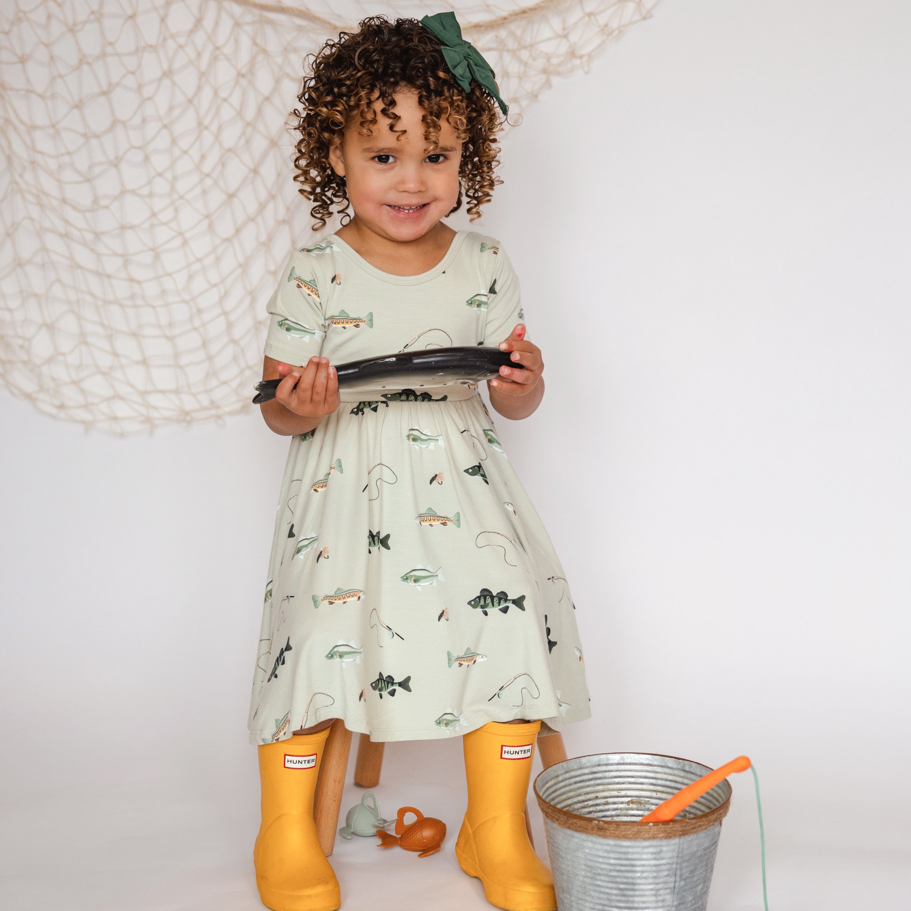 Toddler holding toy fish wearing Twirl Dress in Fishing looking at camera