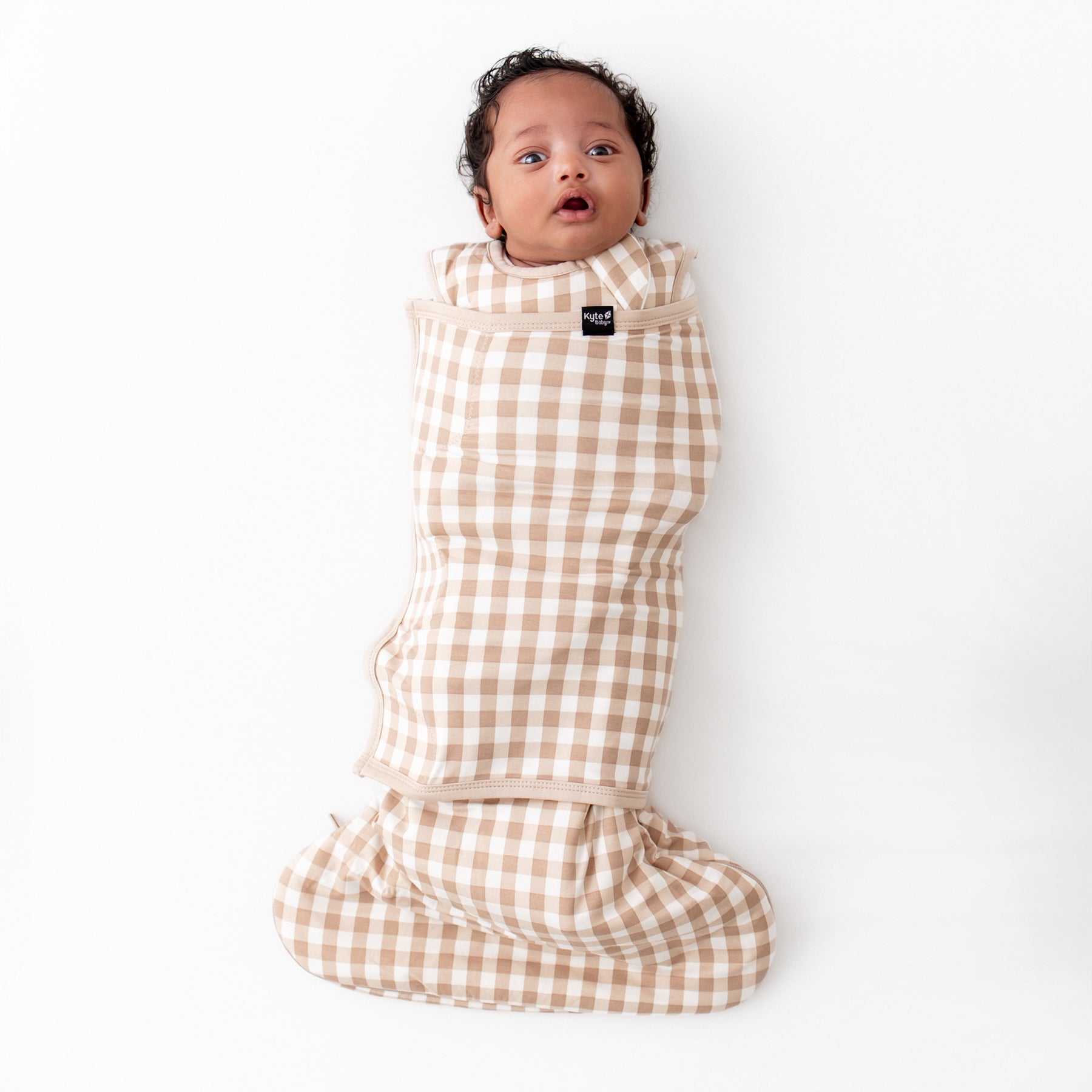 Infant laying down wearing the Sleep Bag Swaddler in Gingham Bisque 1.0