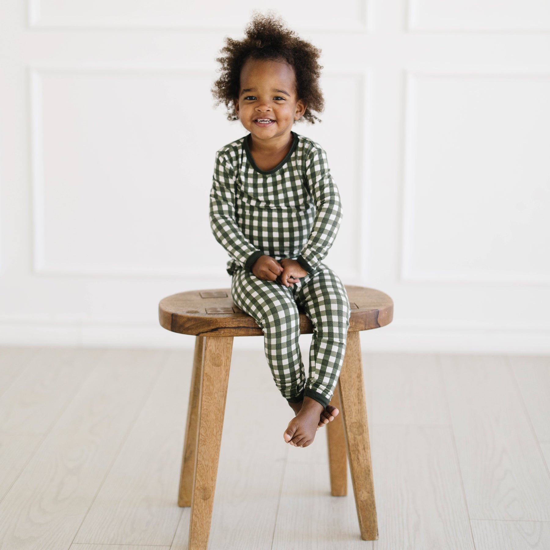 Young toddler sitting on a wooden stool wearing the Long Sleeve Pajamas in Gingham Fir